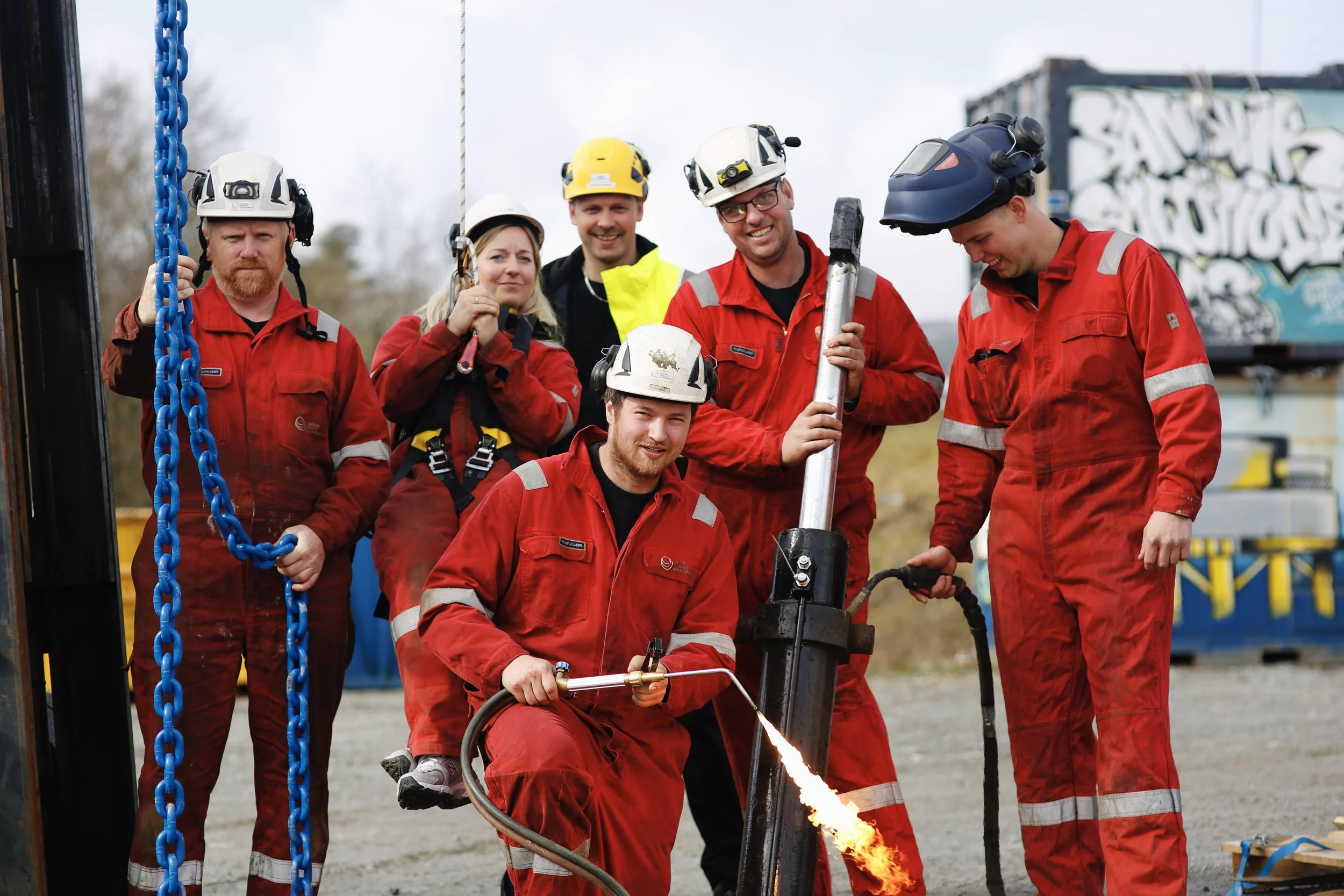 Grupp av redningsarbeidere i røde uniformer og hjelm jobber med å bore i bakken, der en av dem holder en flammekaster, mens de andre observerer.