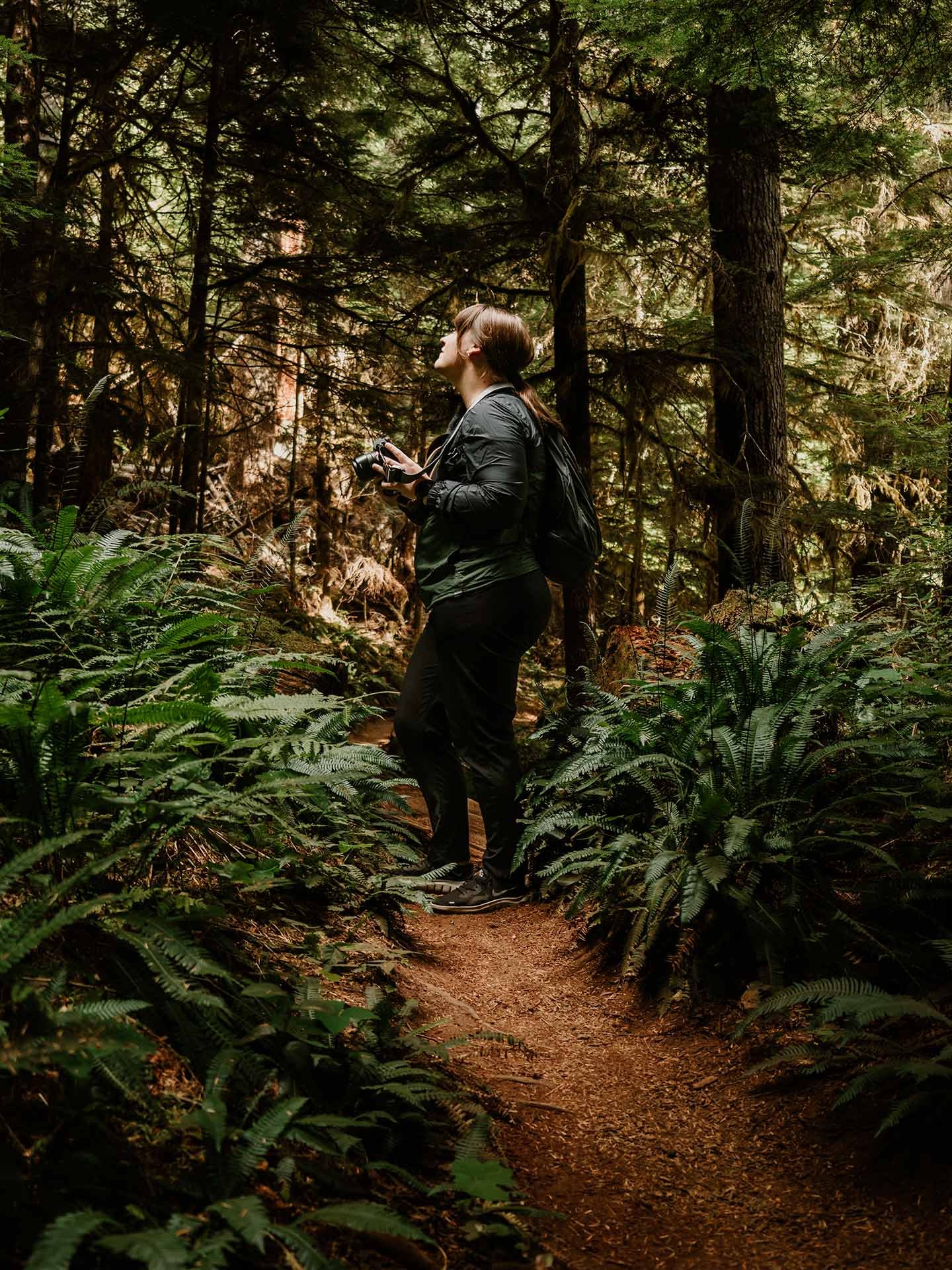 A woman in black outdoor clothing standing on a dirt trail in a dense, green forest while holding a camera.