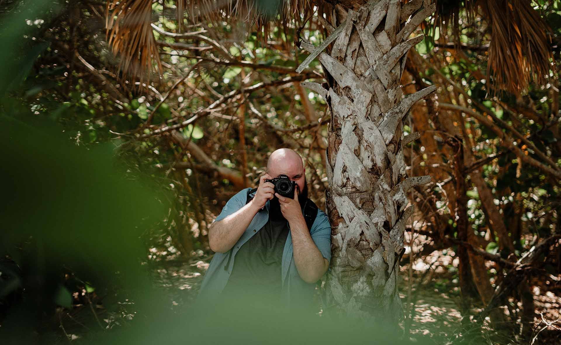 A man taking a photo in a wooded area with large tree trunk and dense foliage.