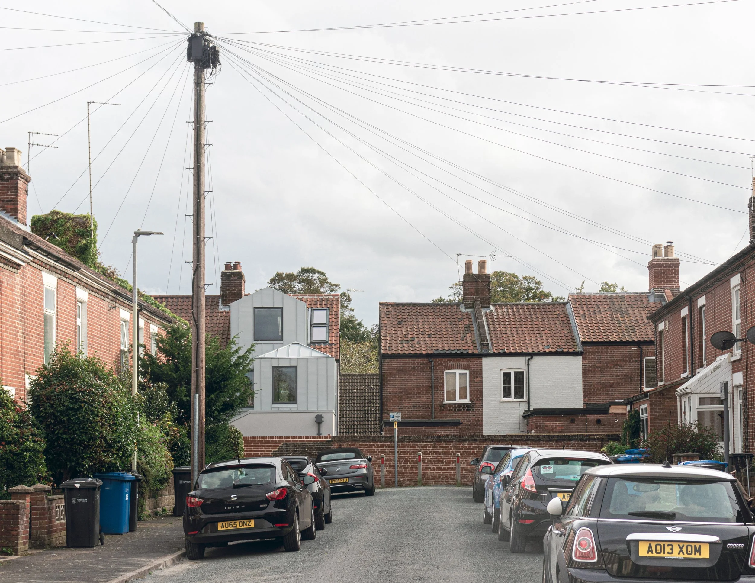 Shortlisted Architect Journal Retrofit Awards - Residential street with parked cars, brick houses, overhead power lines, and a cloudy sky.