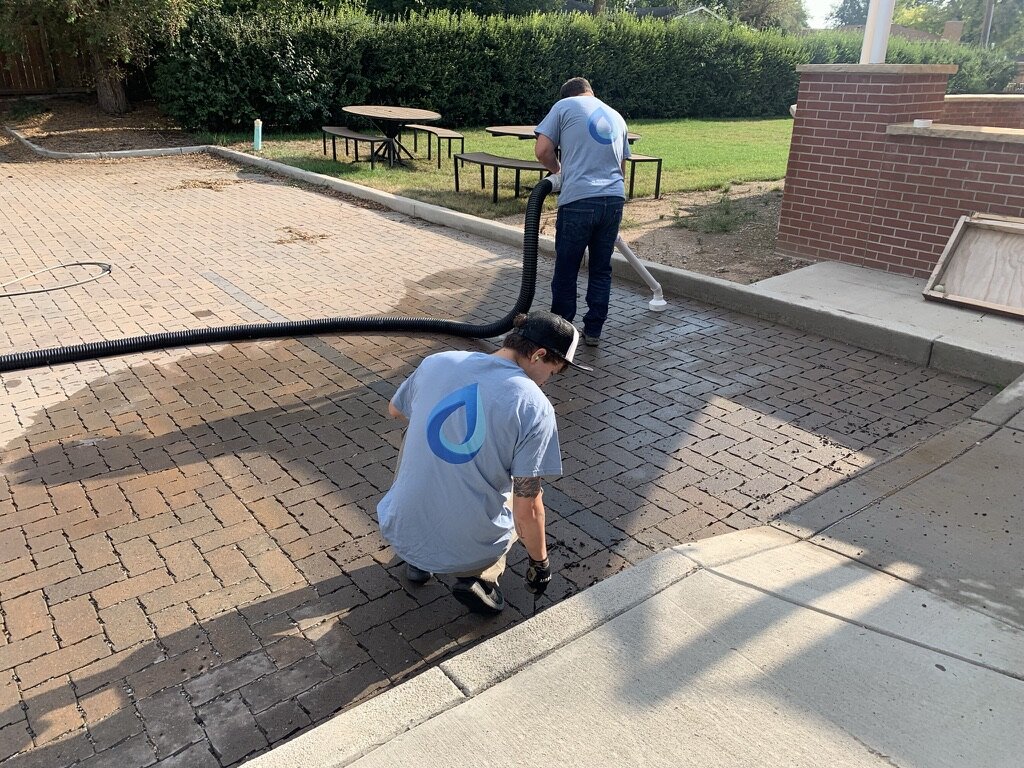 Two workers in blue shirts with a logo are cleaning and pressure washing a brick patio outside a building. One worker is kneeling and scrubbing, while the other is using a hose connected to a pressure washer.