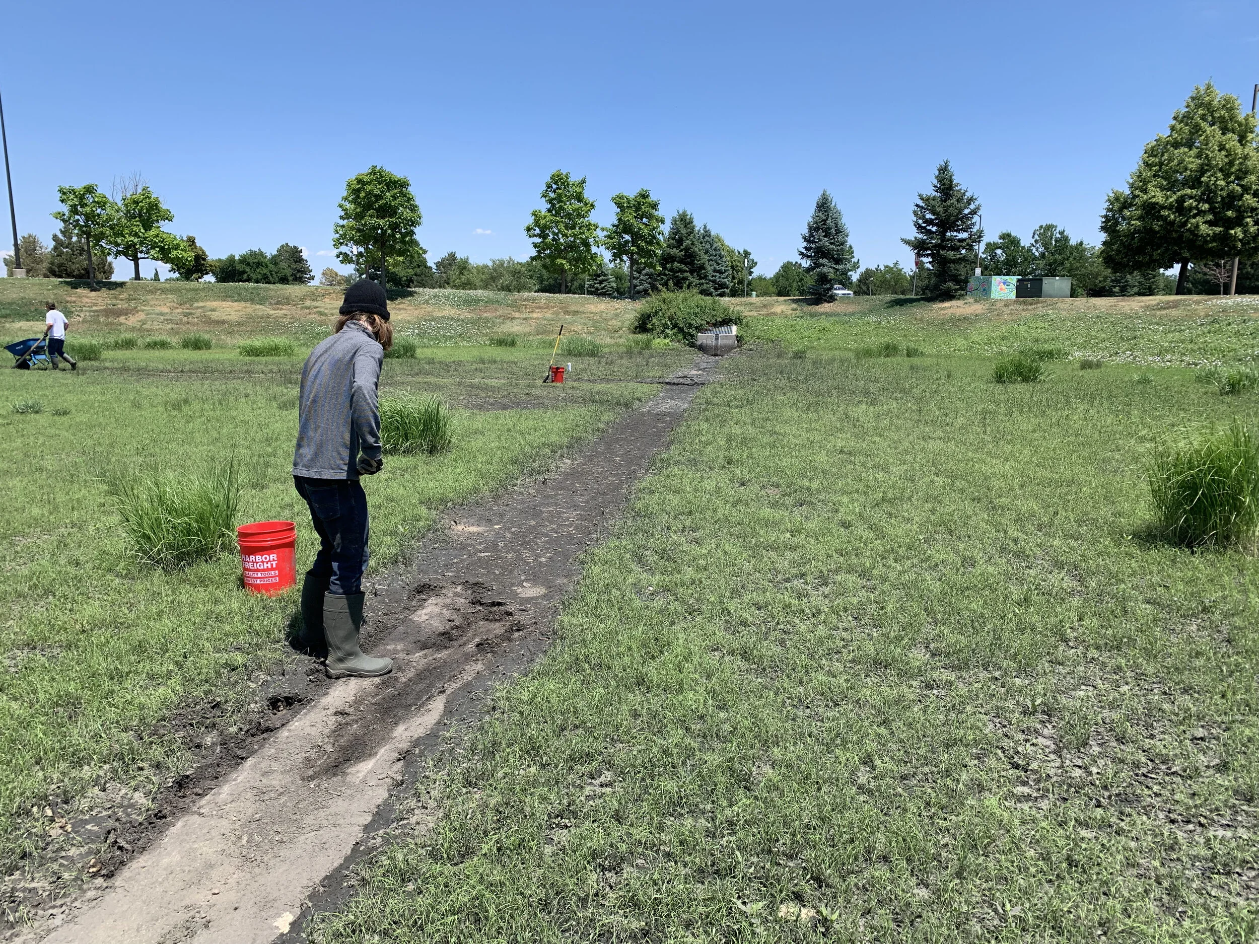 A person standing on a patch of dirt in a grassy field, with another person pushing a wheelbarrow in the background. The field is surrounded by trees and a clear blue sky.