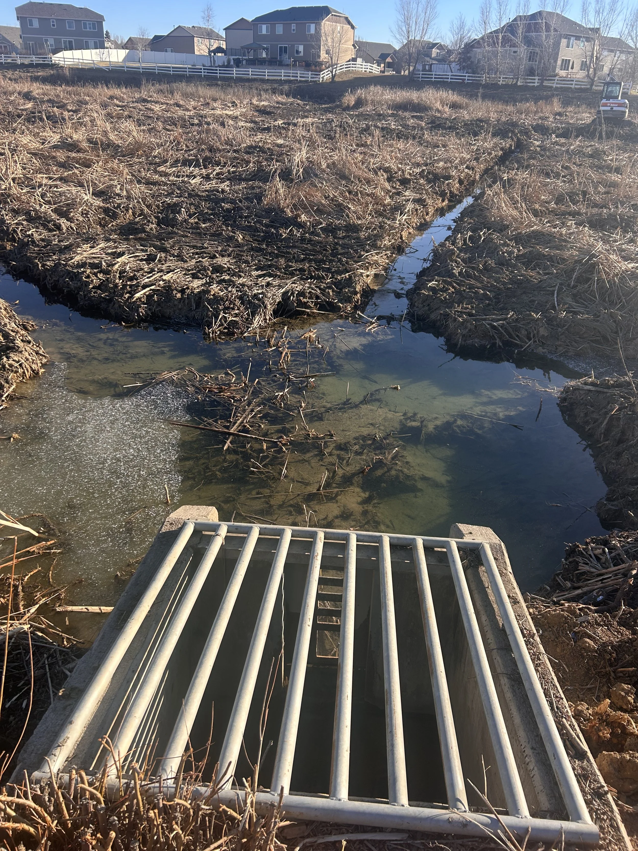 A metal drain grate with a water stream flowing underneath, surrounded by dried grass and mud, with residential houses and a small excavator visible in the background.