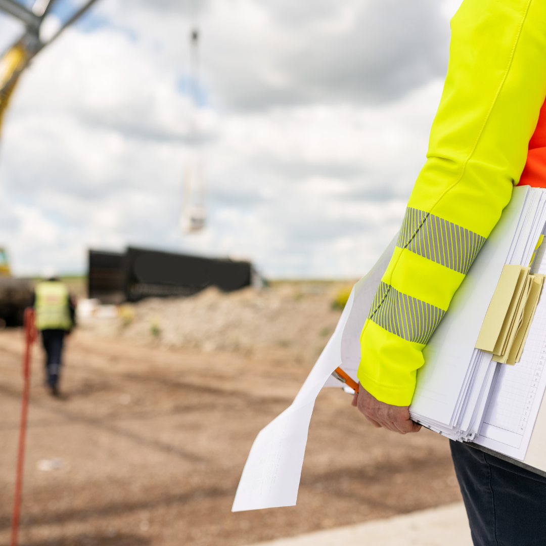Construction worker in a high-visibility yellow jacket holding blueprints at a construction site with a crane in the background.