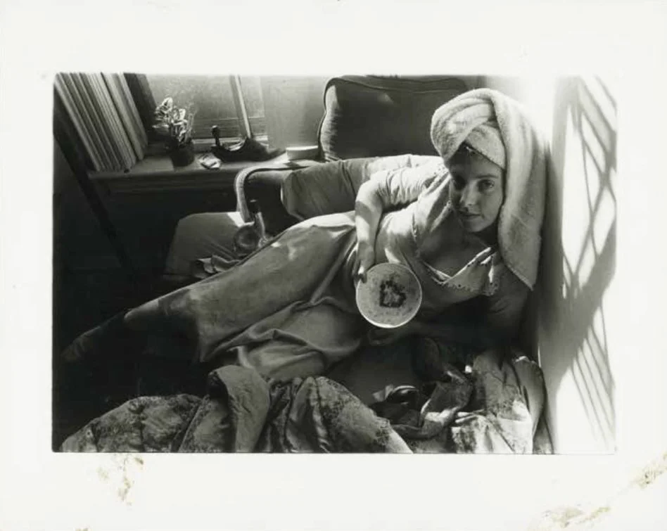 Person lying on a bed with a towel wrapped around their head, holding a bowl, in a room with a window and various objects on the windowsill.