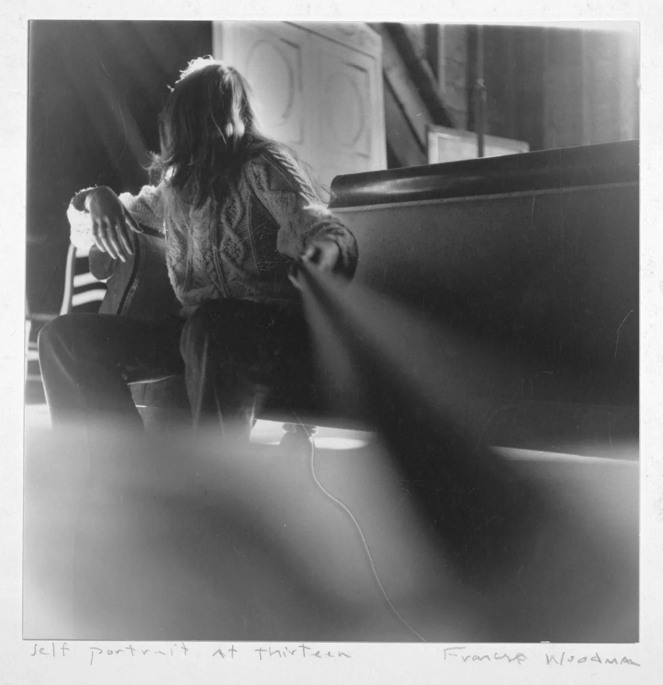 Black and white photo of a young girl sitting on a bench with her arm resting on the back of the bench, in a large indoor space with wooden walls and a door in the background.