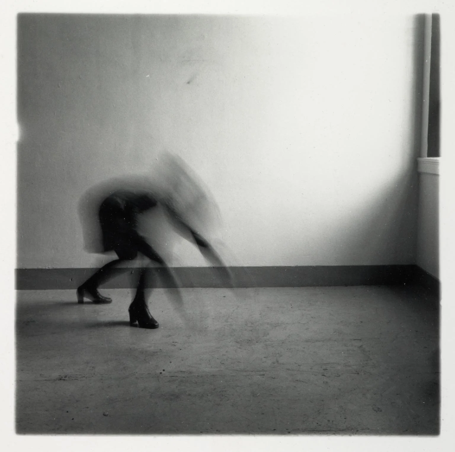 Blurred black-and-white photo of a person in high heels crouching or leaning forward in a room with plain walls and floor.