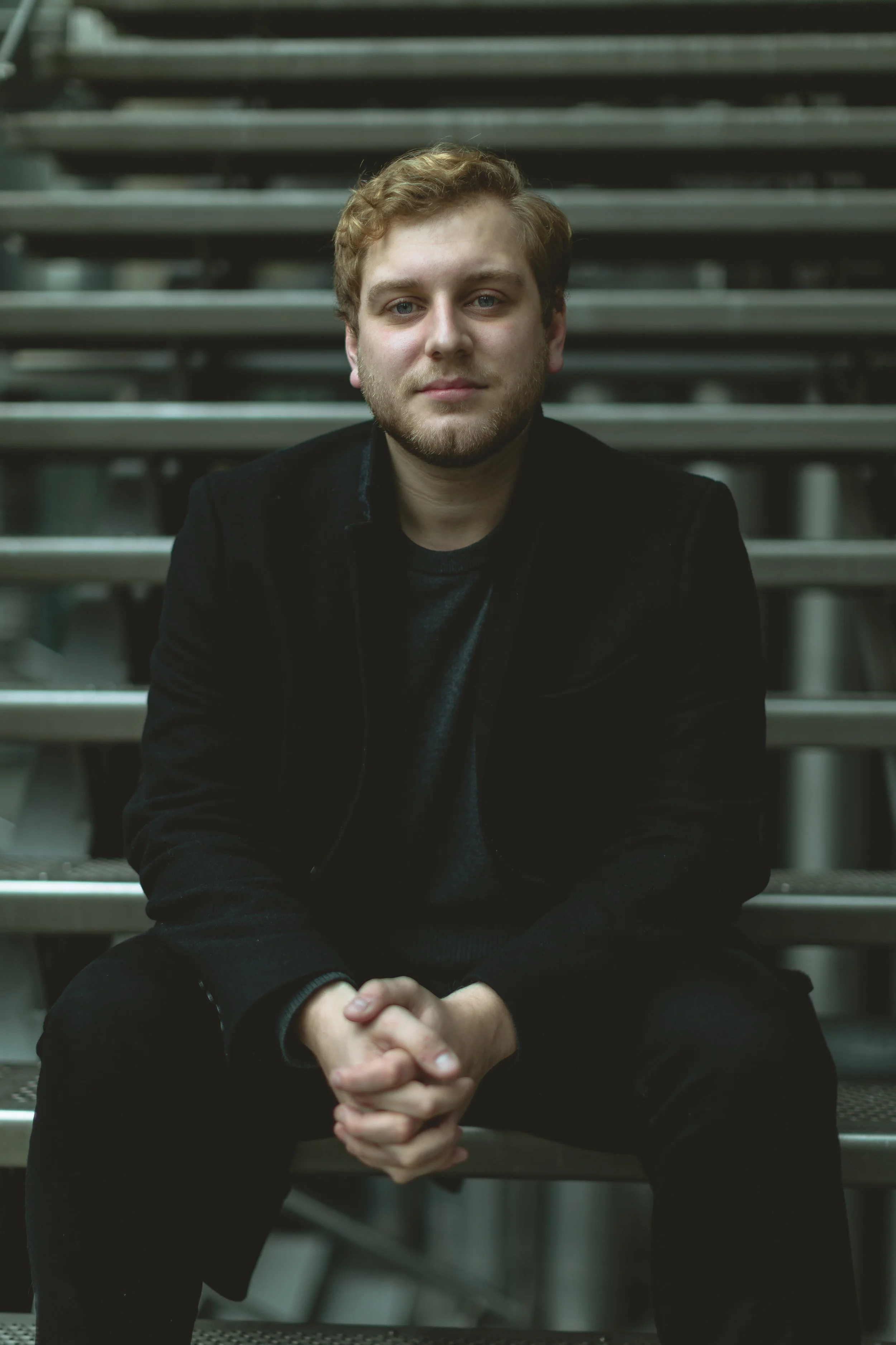 A young man with curly blond hair and light facial hair sitting on metal stairs, wearing a black jacket and dark shirt, looking directly at the camera.