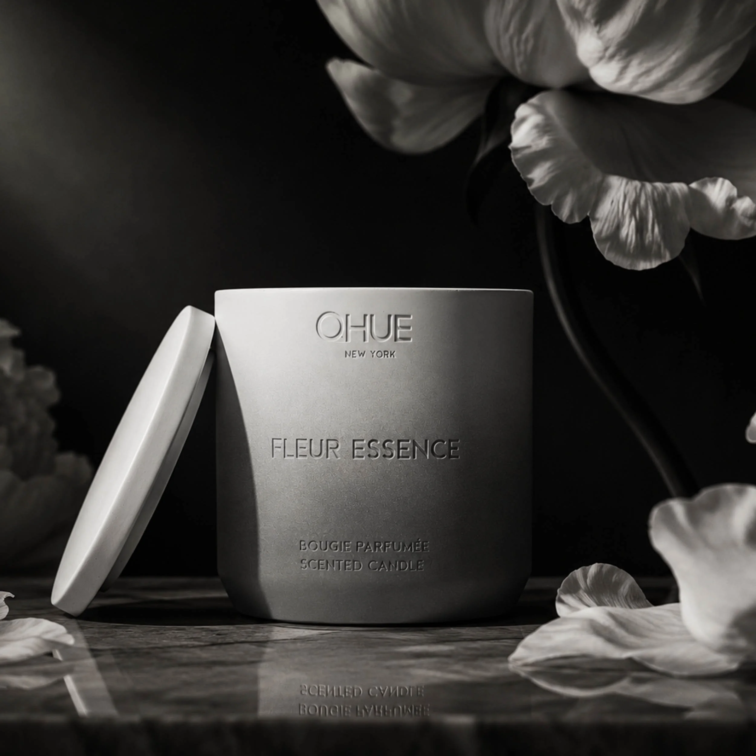 Qhue New York Fleur Essence scented candle in a hand-painted gray cement vessel with lid, styled among white flowers and petals against a dark, elegant backdrop.