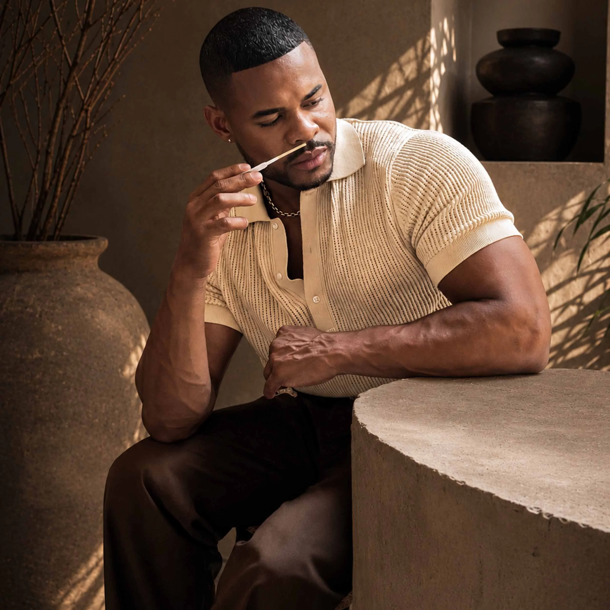 Founder of QHUE New York thoughtfully evaluating a fragrance blotter while seated indoors, wearing a cream knit polo shirt. Soft natural light and lush green foliage create a refined atmosphere, highlighting the craftsmanship and sensory experience