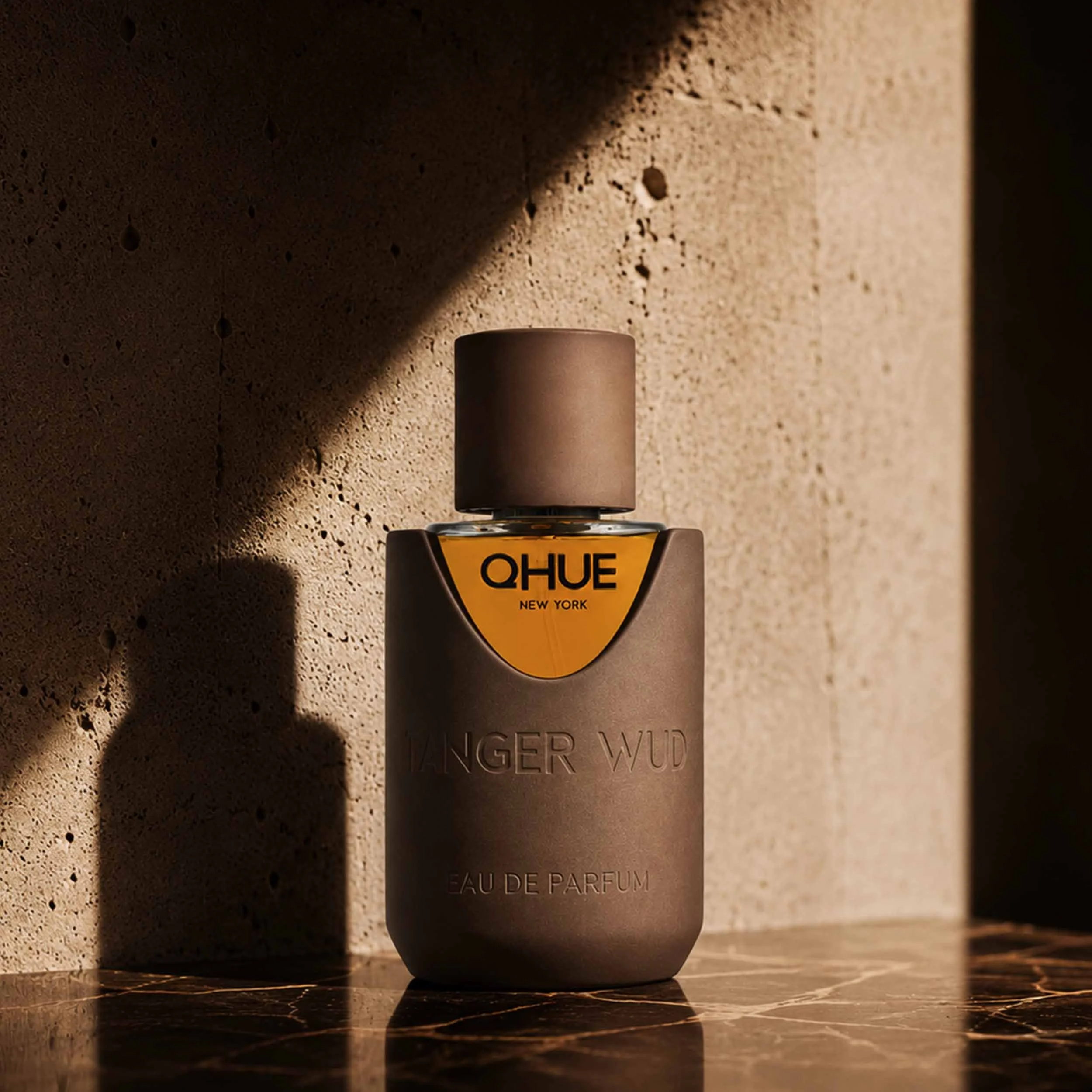 Qhue New York Tanger Wud Eau de Parfum in a hand-painted brown bottle with golden fragrance, displayed against a warm architectural backdrop.