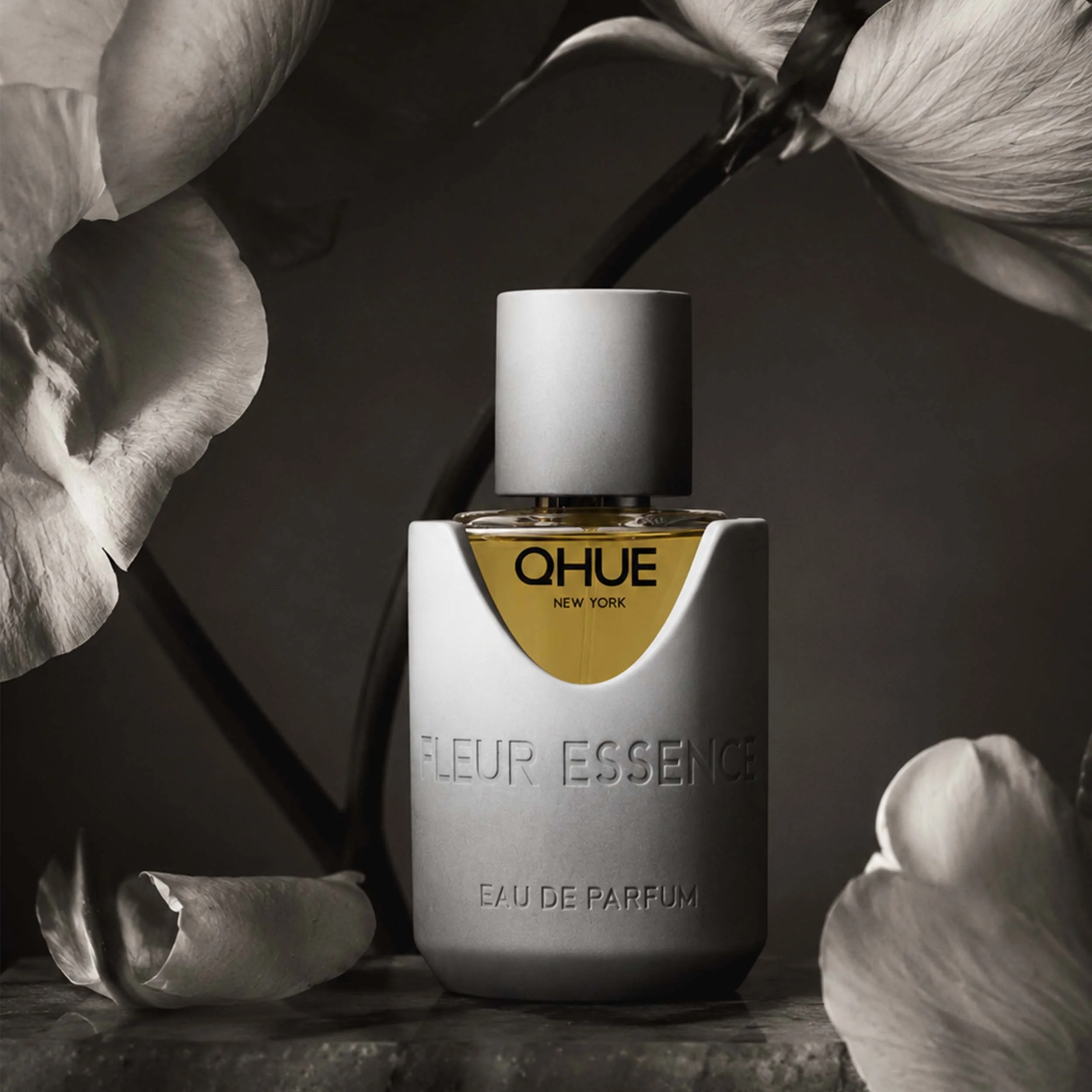 Qhue New York Fleur Essence Eau de Parfum in a hand-painted gray and white bottle with golden fragrance, surrounded by white flowers in a luxurious floral setting.