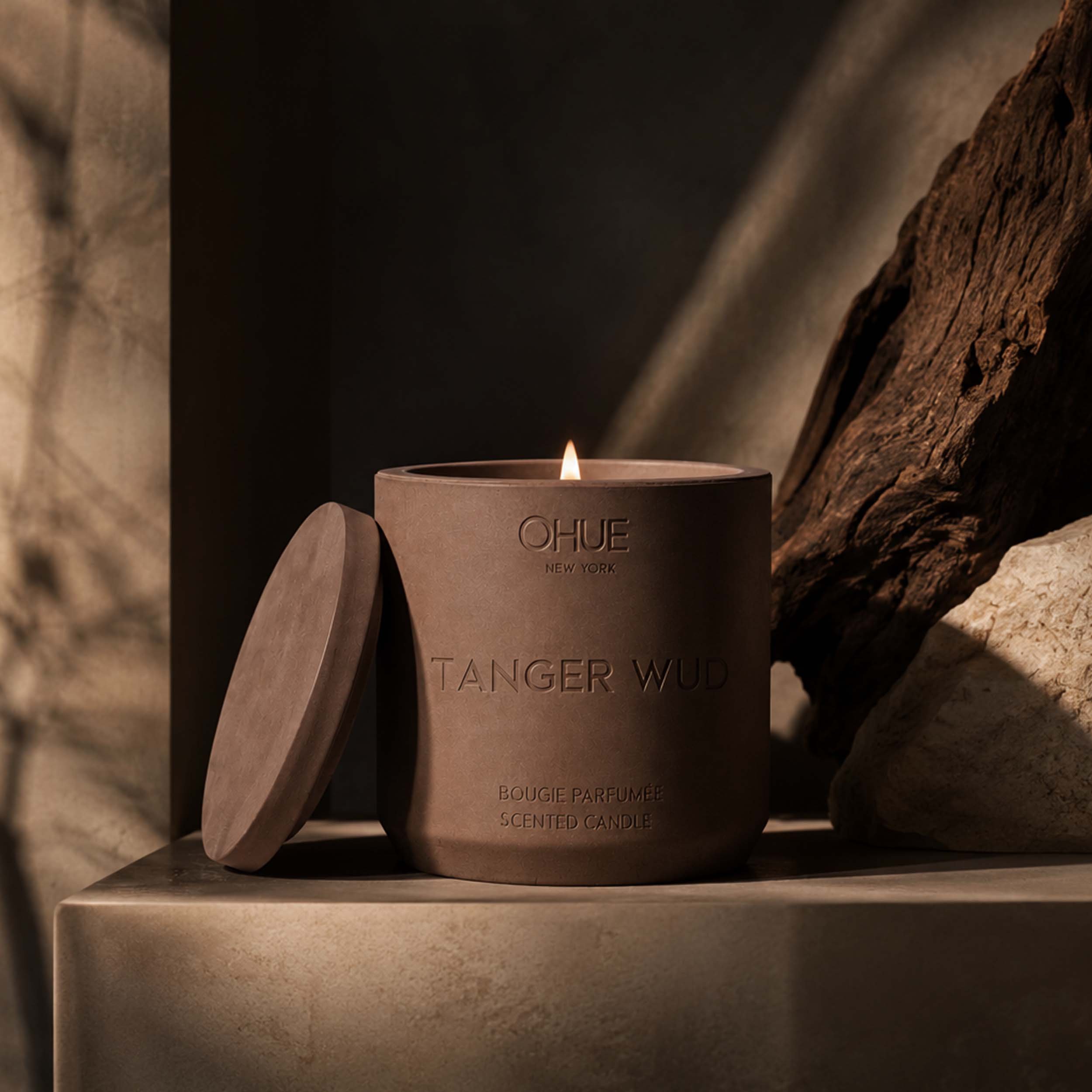 Qhue New York Tanger Wud scented candle in a hand-painted brown cement vessel with lit flame, styled with natural wood and stone in warm lighting.