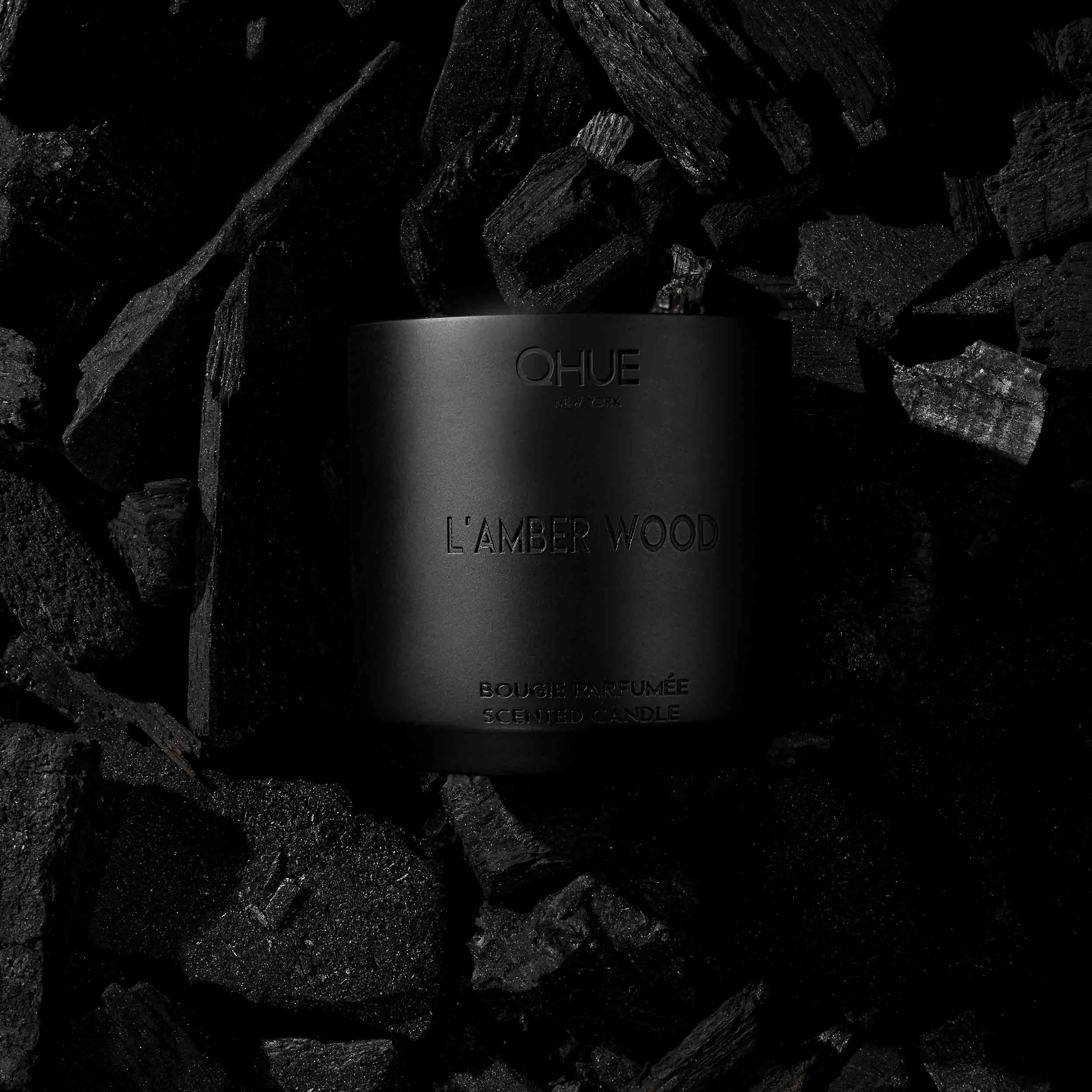 Qhue New York L’Amber Wood scented candle surrounded by black charcoal.