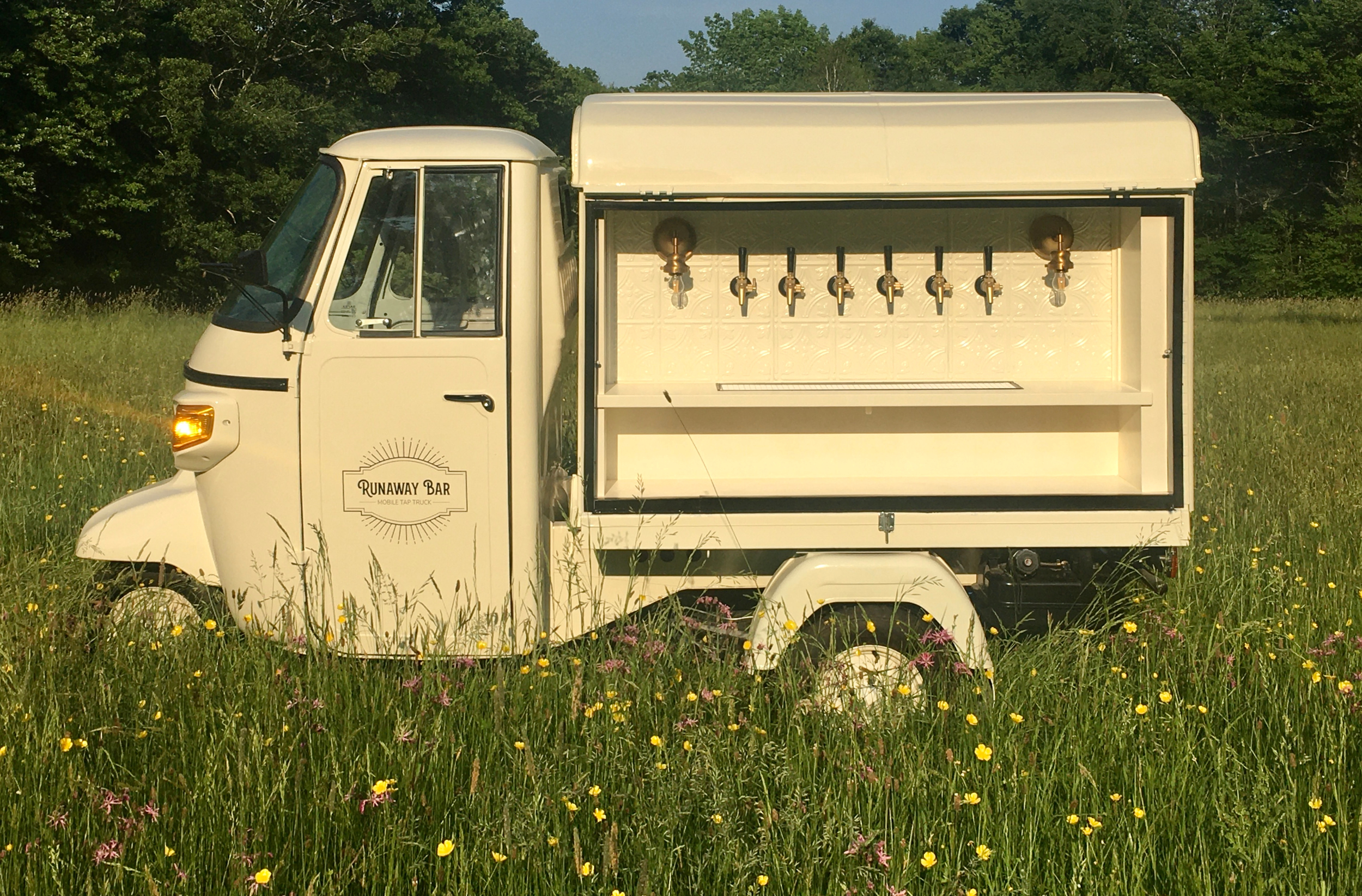 A white mobile tap truck labeled 'Runaway Bar' with multiple beer taps on a side panel, parked in a grassy field with wildflowers, surrounded by trees under a blue sky.