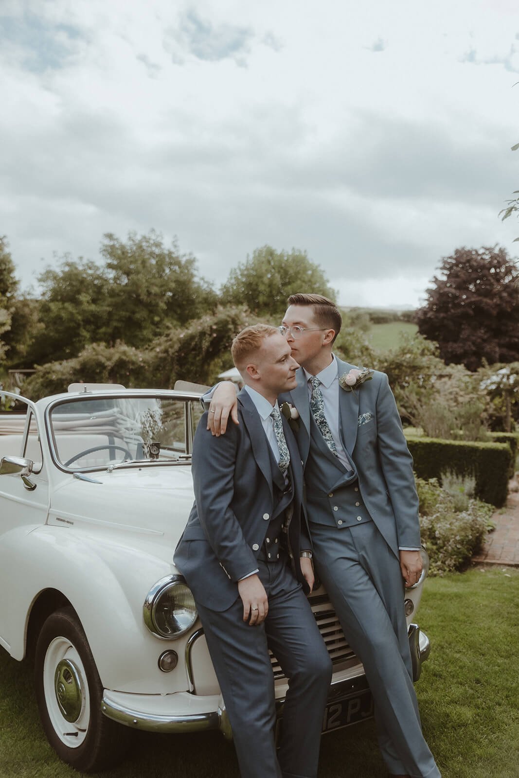 Two grooms in matching suits and floral ties are sharing an embrace and a kiss outside, leaning against a vintage white car on a grassy lawn with trees and a cloudy sky in the background.