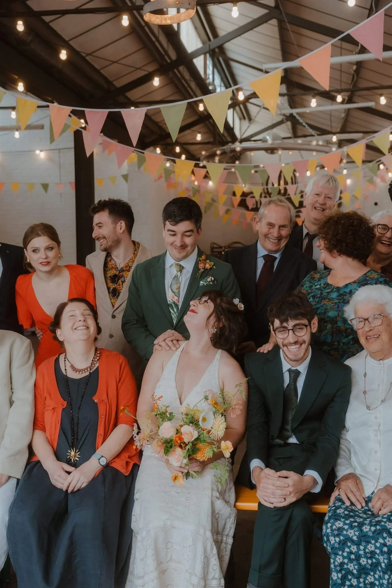 A group of people at a celebration, gathered indoors under hanging string lights with colorful bunting flags overhead. The group includes a bride holding a bouquet, surrounded by family and friends.