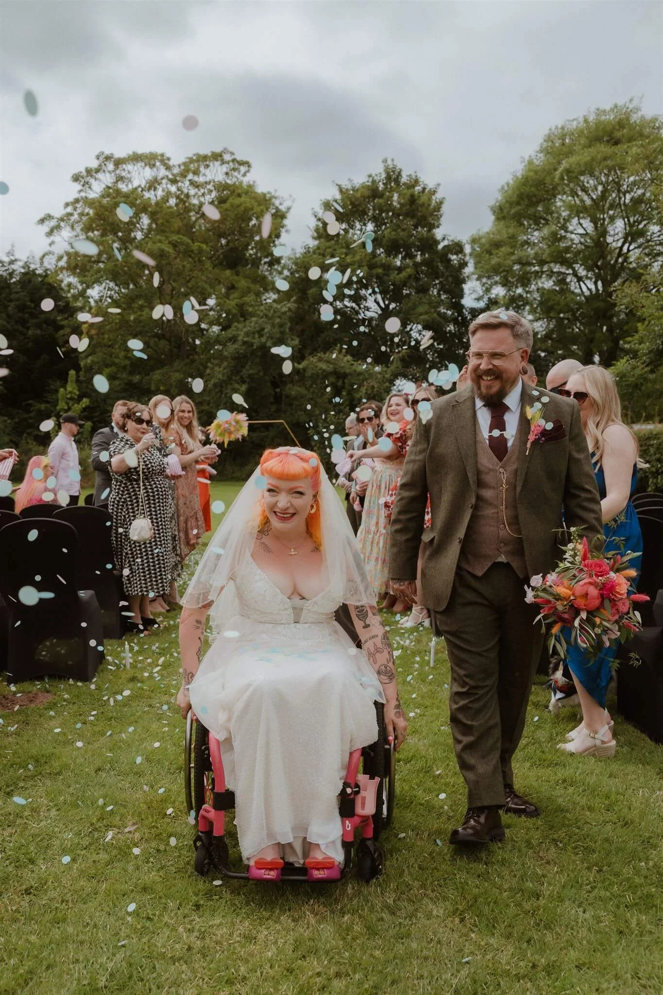A bride in a wedding dress and veil is smiling and coming down the aisle in a wheelchair at her wedding, and the groom holding a bouquet of flowers. Guests are celebrating and throwing confetti in the background, outdoors with trees and a cloudy sky.