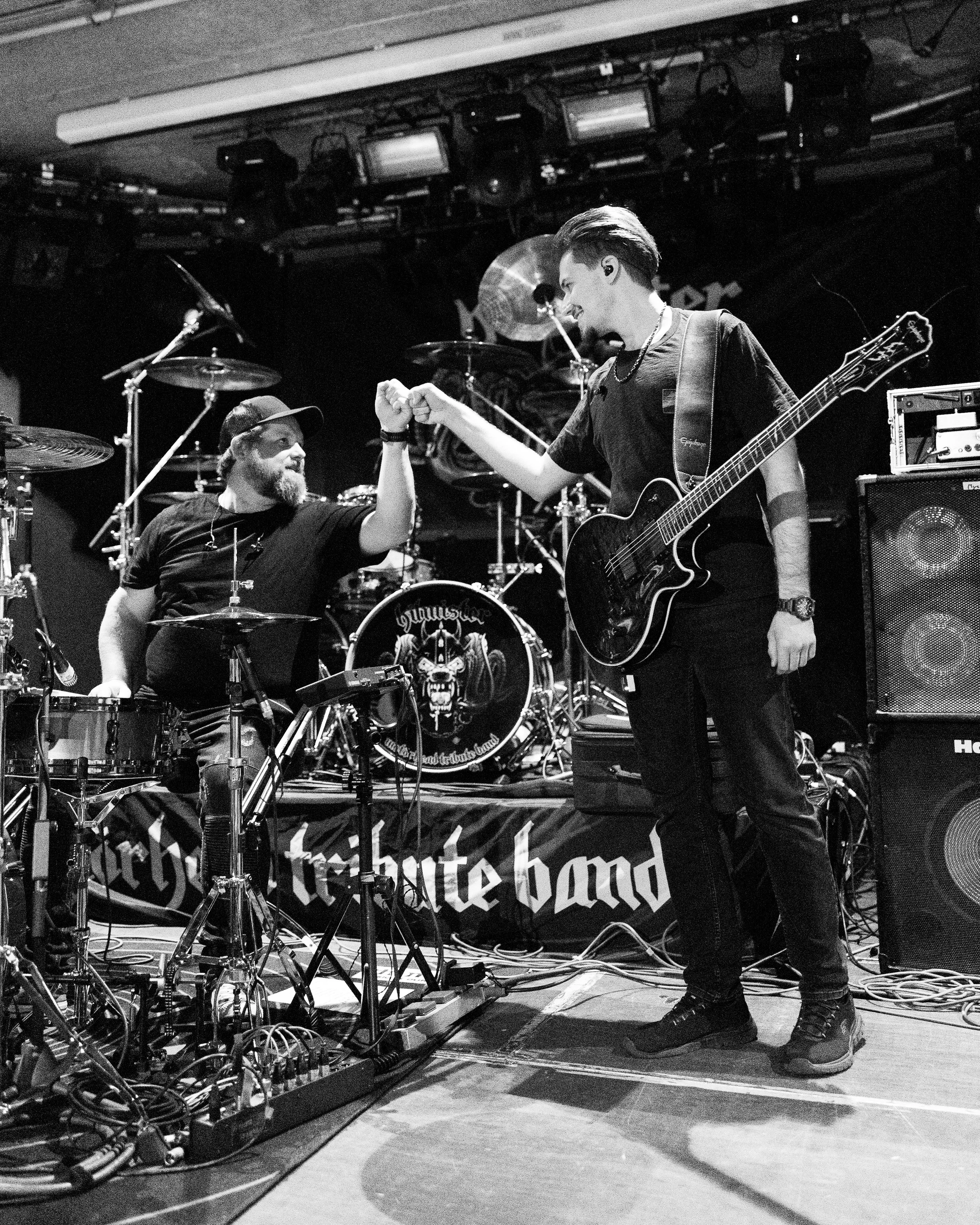 Zwei Musiker auf einer Bühne, die sich die Faust geben, eines mit Schlagzeug und das andere mit Gitarre, in einem Schwarz-Weiß-Foto.