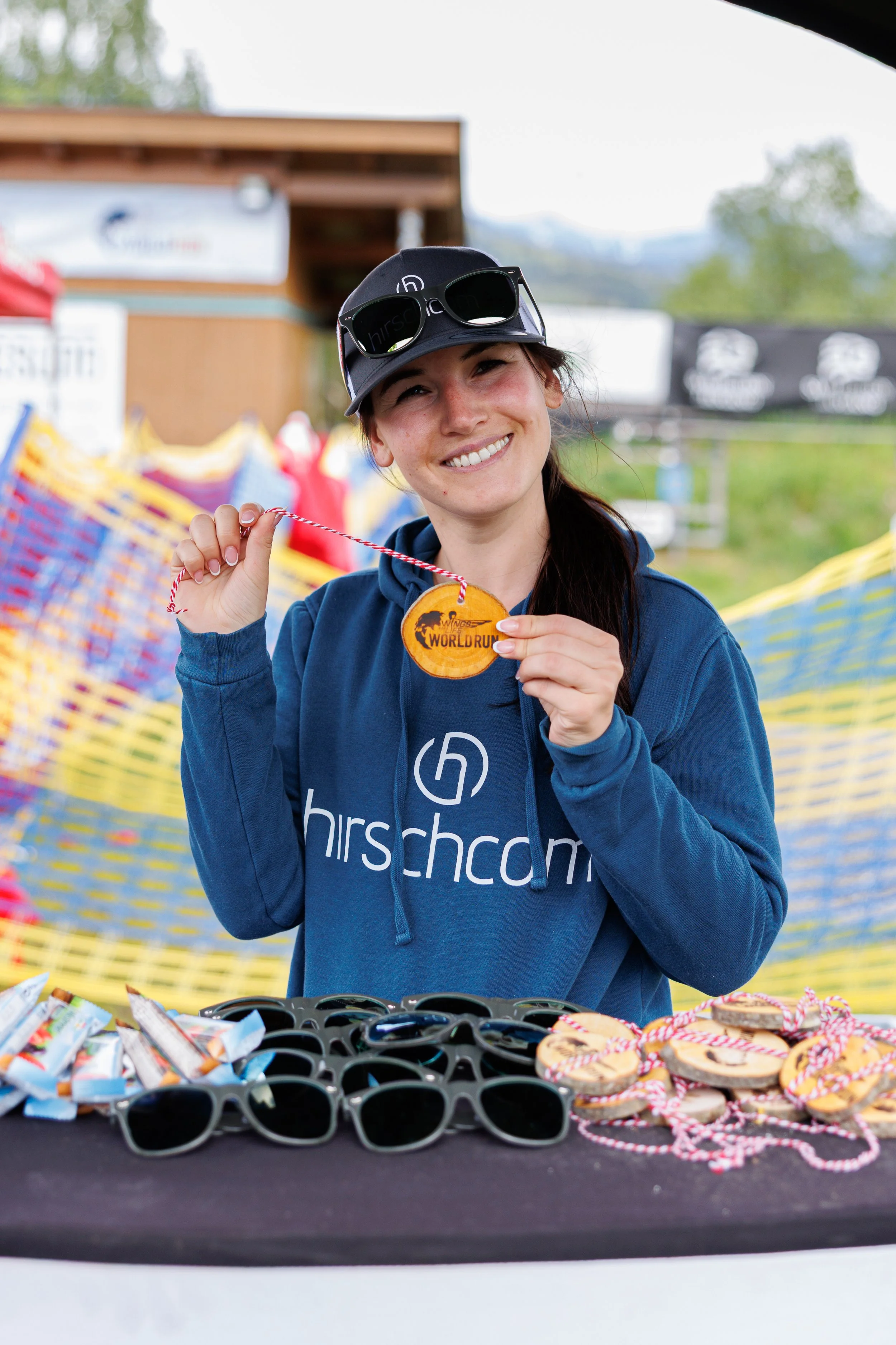 Junge Frau zeigt eine bronze Medaille bei einem Sportevent, mit mehreren Sonnenbrillen und Medaillen auf einem Tisch vor ihr.