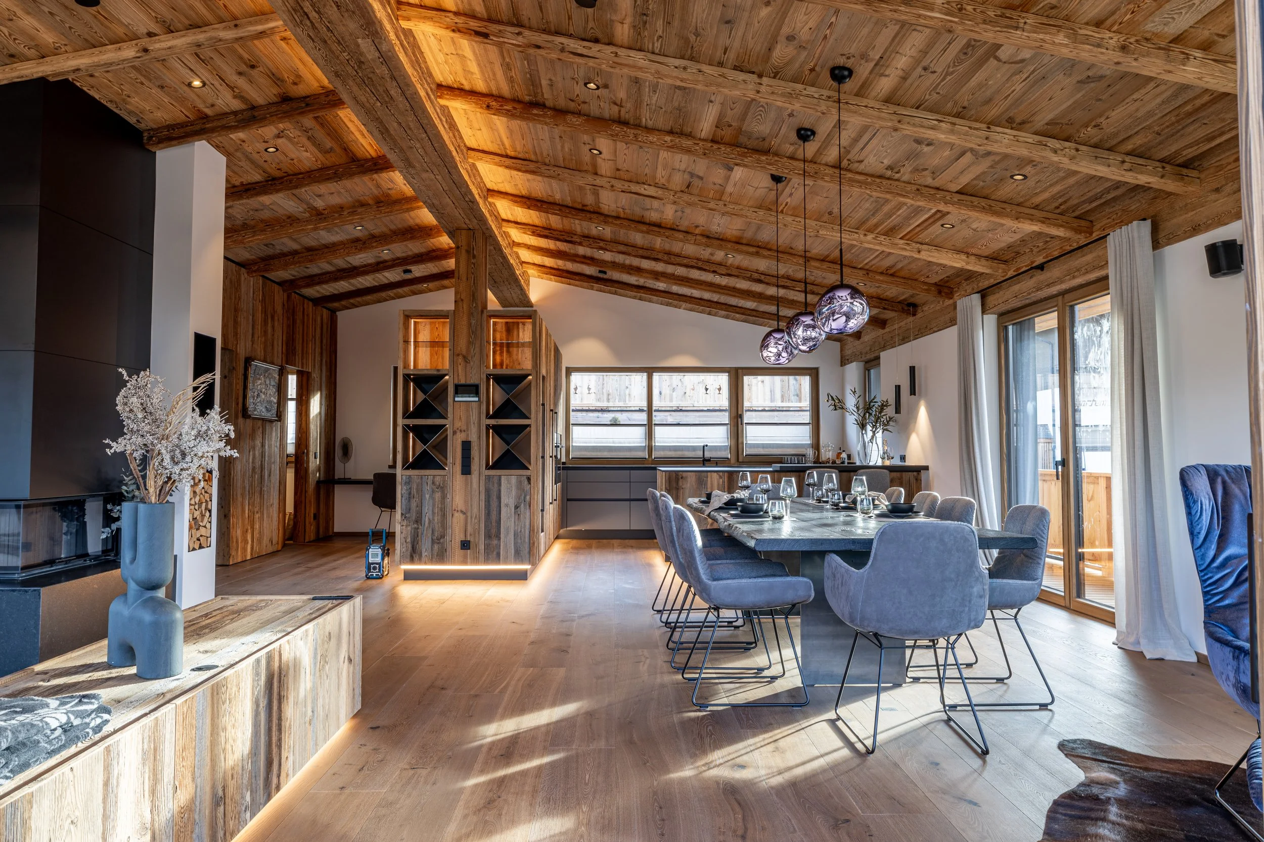 Ein modernes Esszimmer mit Holzdecke, Holzfußboden und großen Fenstern. Es gibt einen langen Tisch mit sechs bequemen Stühlen, einer modernen, hängenden Beleuchtung und Dekorationen wie Vasen und Pflanzen.