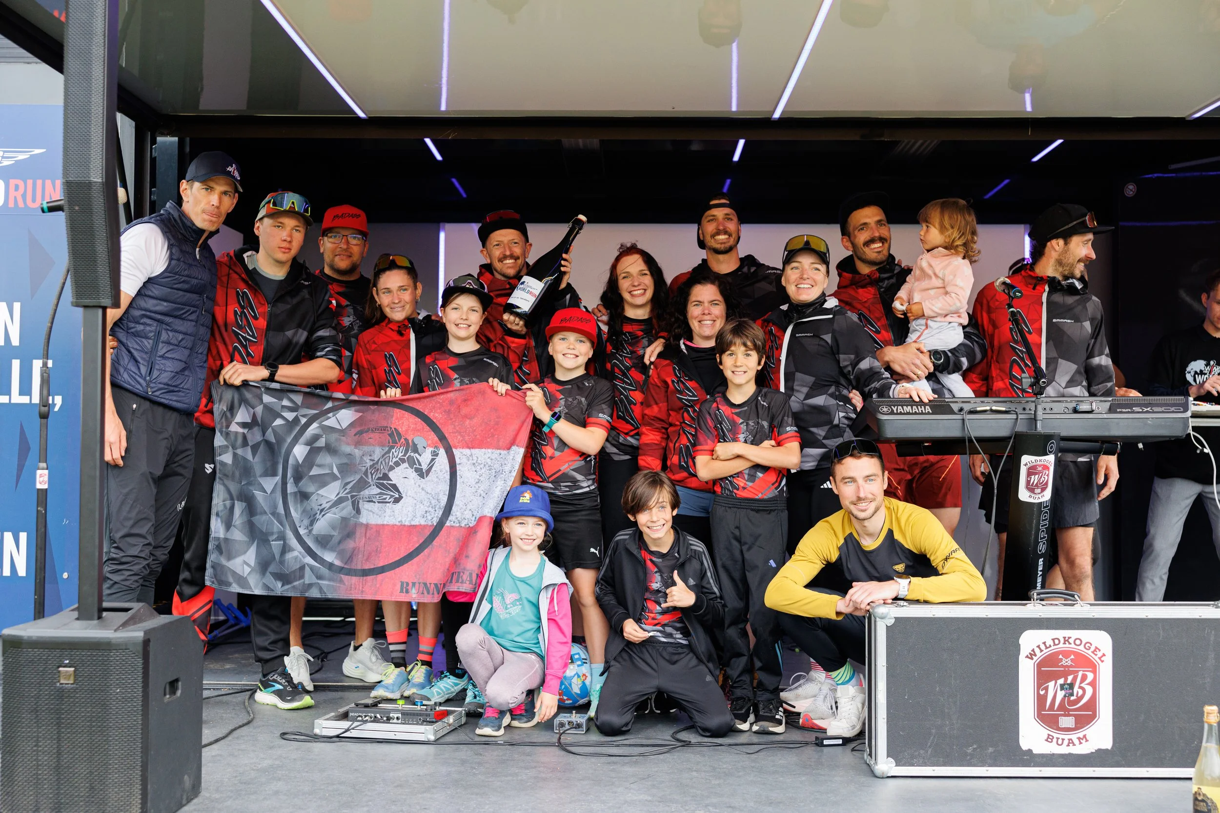 Gruppe von Menschen in Teamkleidung auf einer Bühne, einige halten ein Flagge mit einem Pantherzeichen, jubeln und lächeln, im Hintergrund LED-Lichtinstallationen, einige Kinder, Getränke, und technische Ausrüstung.