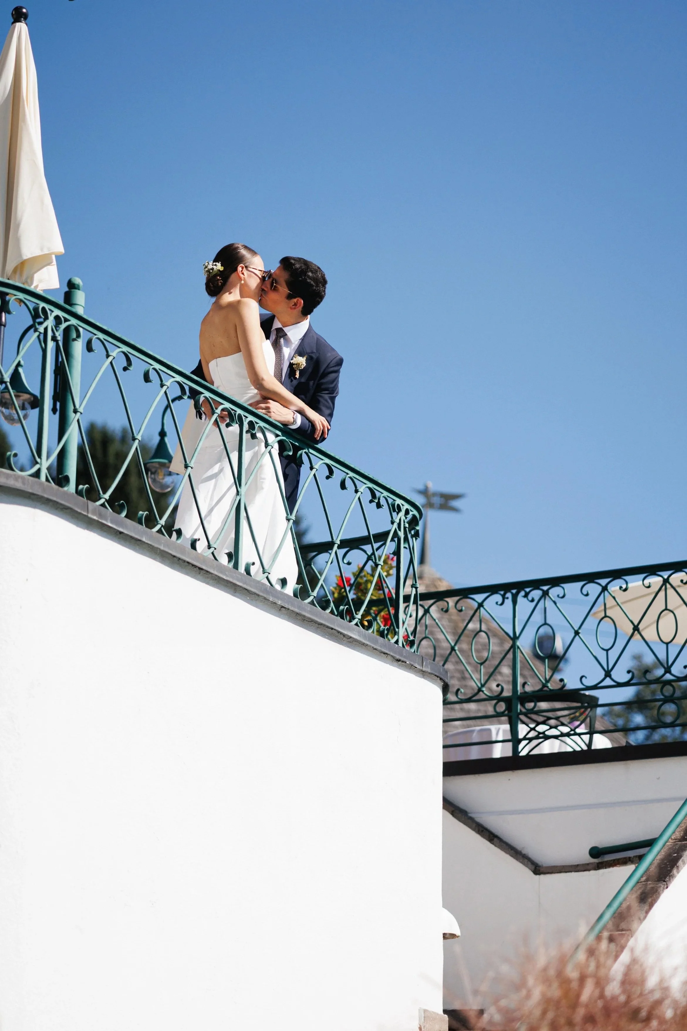 Ein Paar bei einer Hochzeitszeremonie, das sich auf einem Balkon küsst, mit klarem blauen Himmel im Hintergrund.