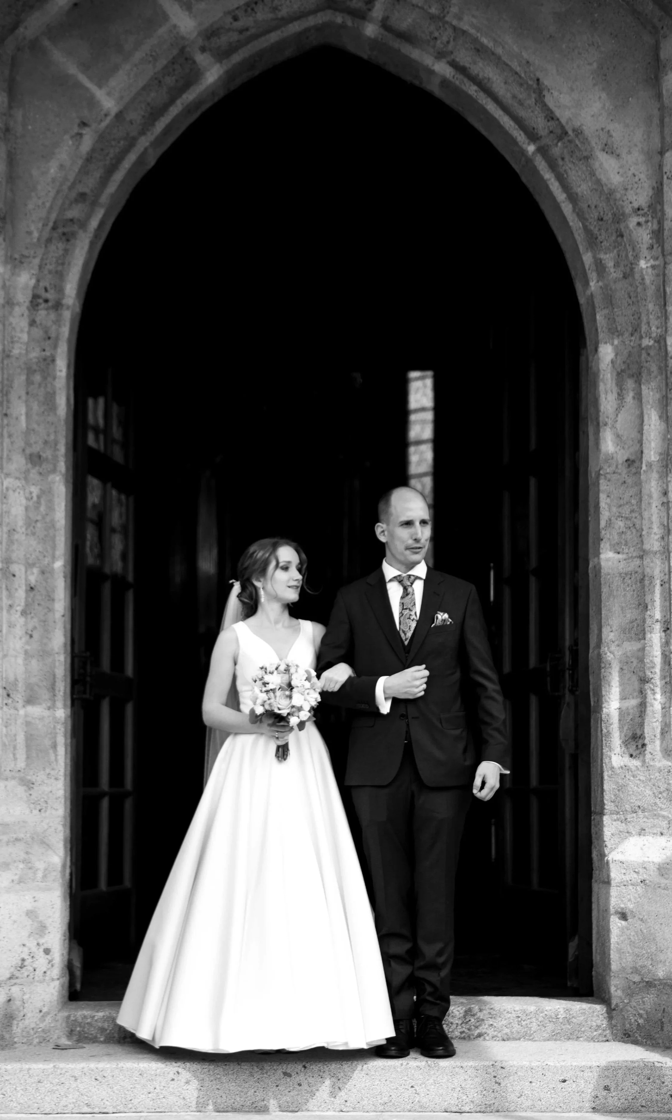 Braut in Hochzeitskleid mit Brautstrauß und Bräutigam in Anzug, die aus einer Kirche oder historicem Gebäude aus schwarzem Stein treten.