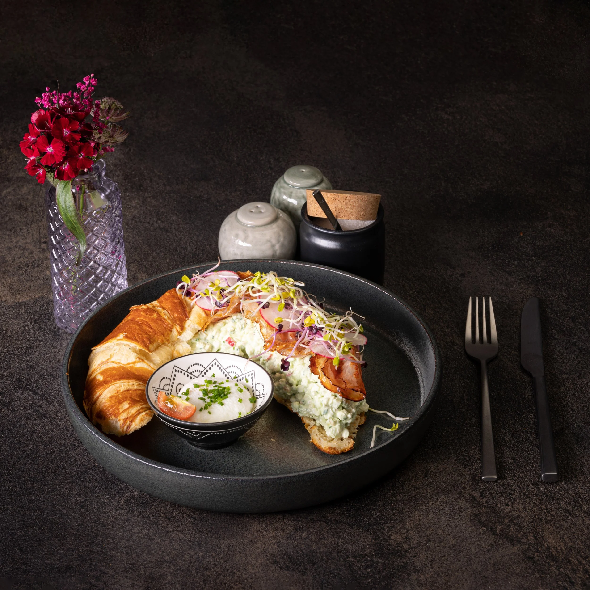 Ein dunkler Teller mit Croissant, Kartoffelsalat, Radieschen, Sprossen und einem kleinen Schälchen mit Sauce. Im Hintergrund eine Vase mit roten Blumen, Salz- und Pfefferstreuer sowie eine schwarze Gewürzbox. Besteck liegt rechts auf dem Tisch.