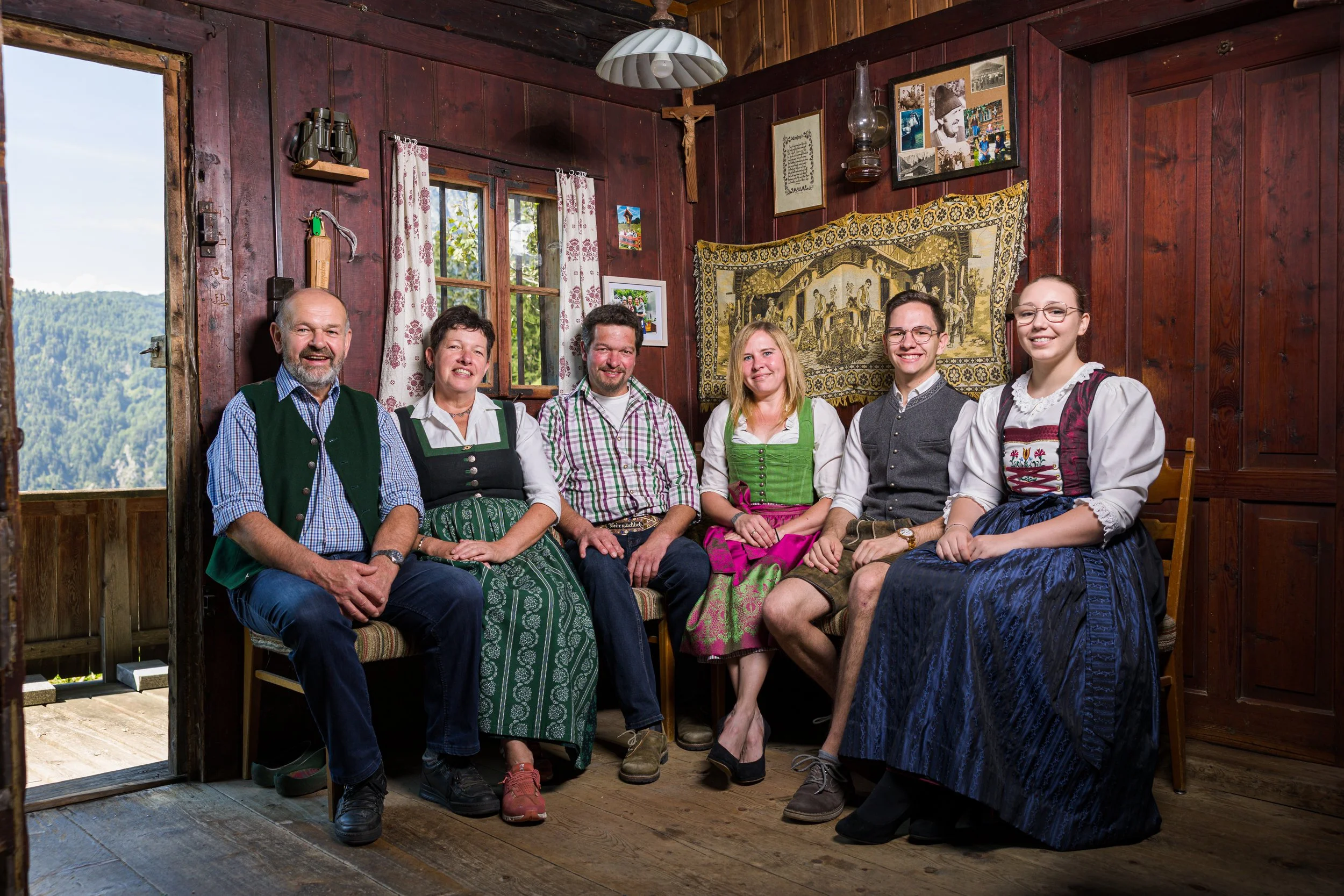 Eine Gruppe von sieben Personen in traditionellen bayerischen Trachten sitzt auf einer Bank in einer rustikalen, holzvertäfelten Hütte, mit Blick auf die Berge im Hintergrund.