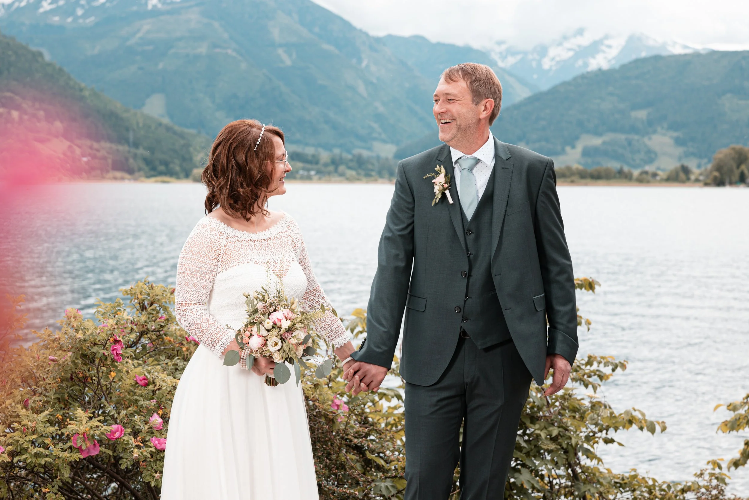 Ein frisch verheiratetes Paar hält Händchen an einem See mit Bergen im Hintergrund, beide lachen glücklich bei ihrer Hochzeit.