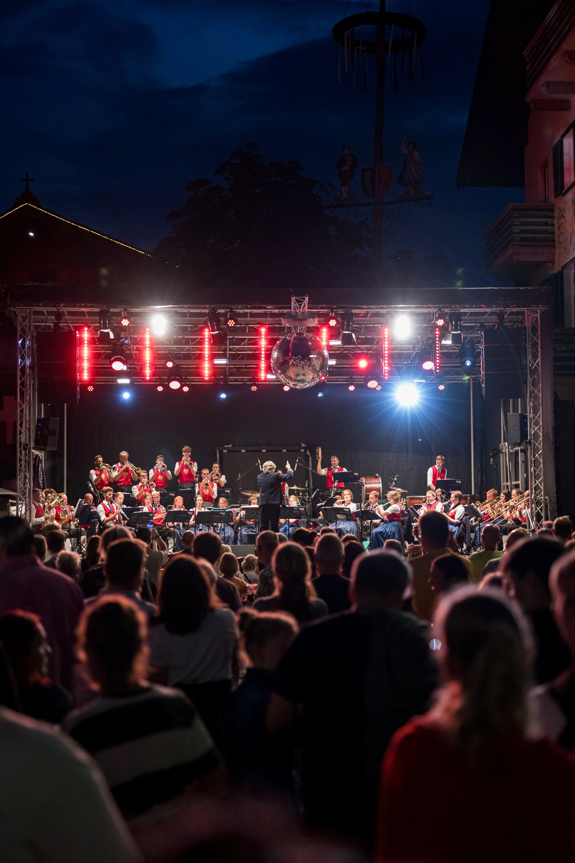 Ein Orchester spielt auf einer Bühne während eines Open-Air-Konzerts bei Nacht, mit einem Publikum im Vordergrund.