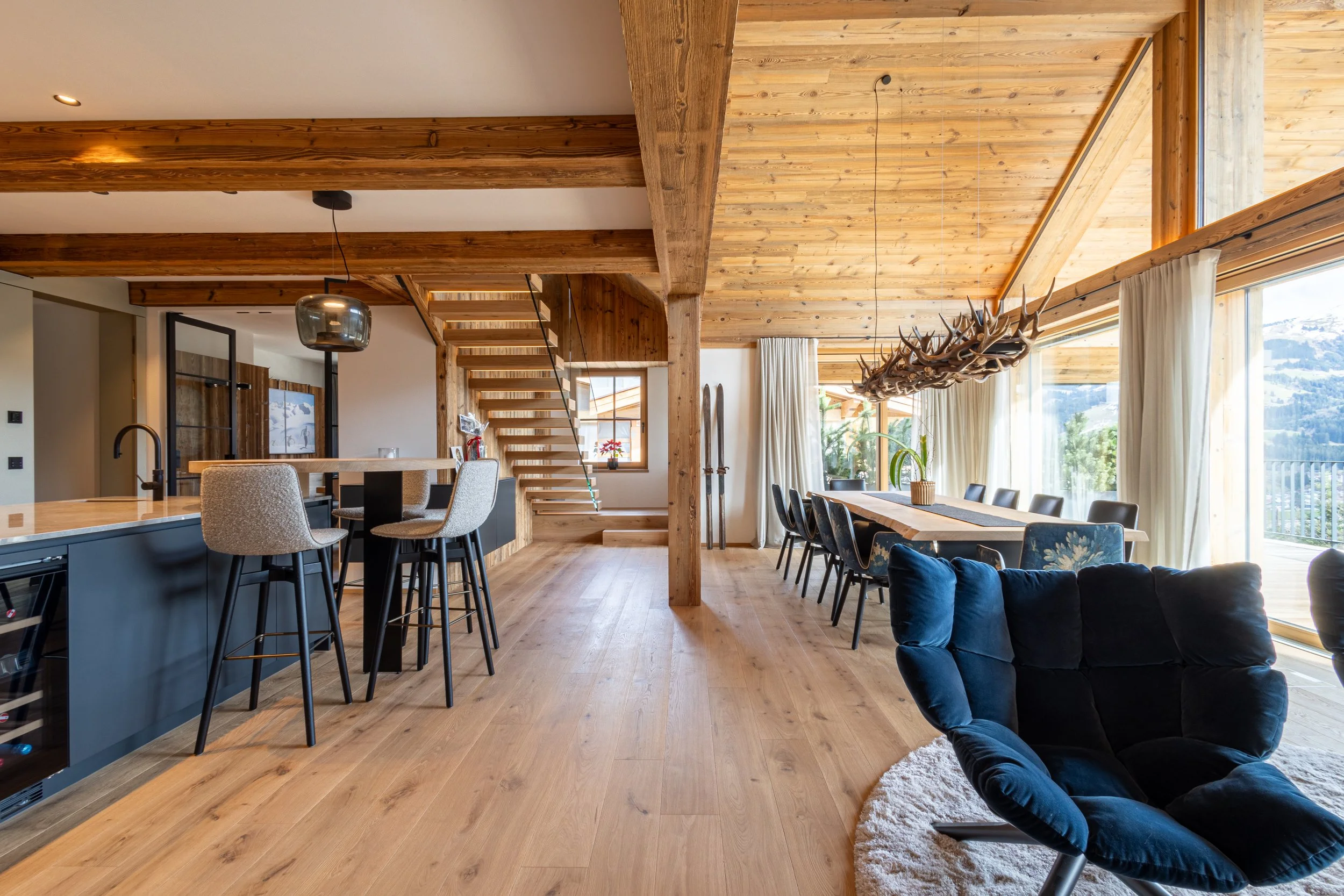 Offener Wohn- und Essbereich mit Holzdecke, großem Esstisch, Fenster mit Blick auf Berge, moderne Küche mit Bartheke, dunkle Sitze, antler-Leuchter, Holzfußboden