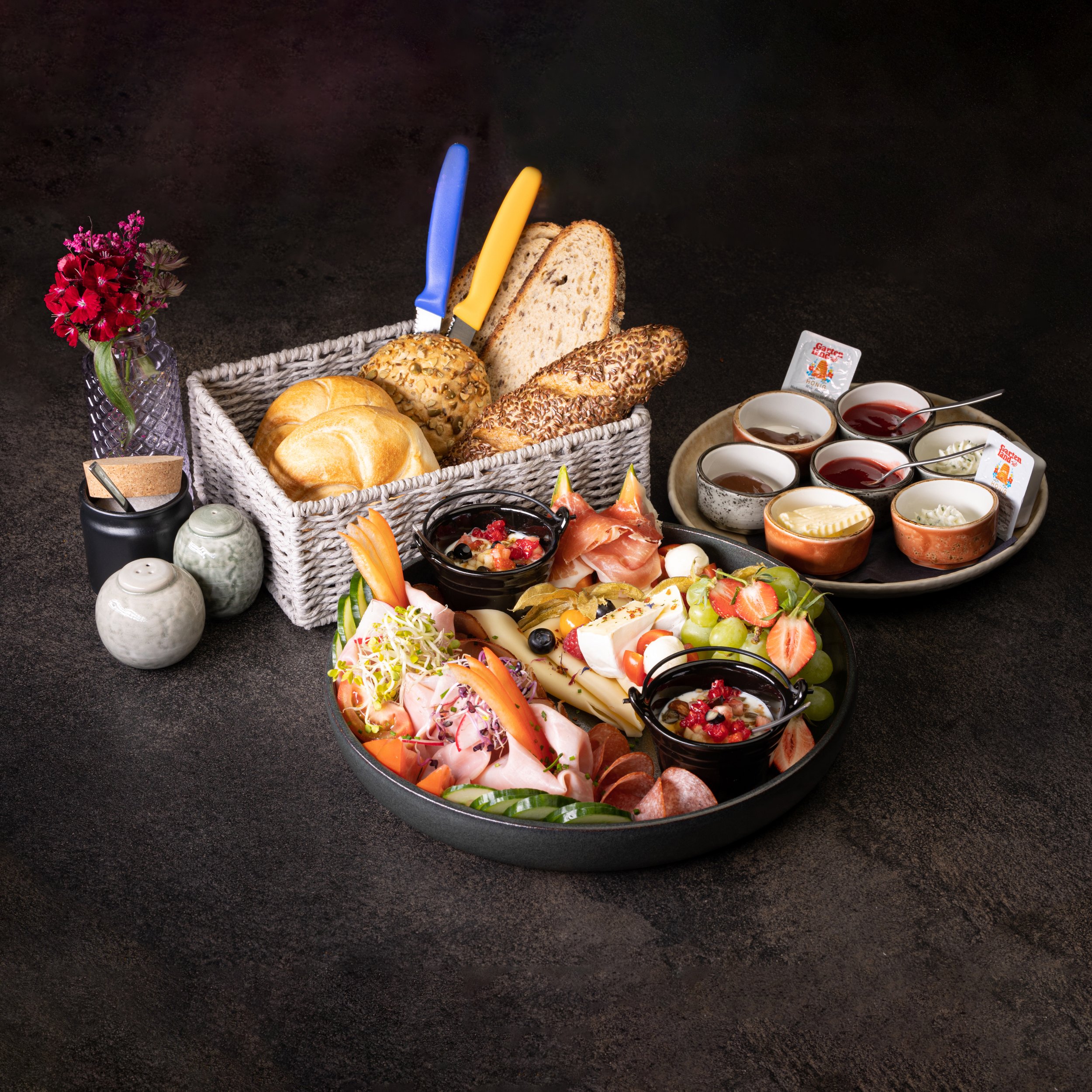 Ein Frühstücksteller mit verschiedenen Wurstsorten, Käse, frischen Früchten, Sektgläser und Marmeladen. Neben einem Korb mit Brötchen, Semmeln und Baguette. Ein kleines Vase mit roten Blumen ist ebenfalls zu sehen.