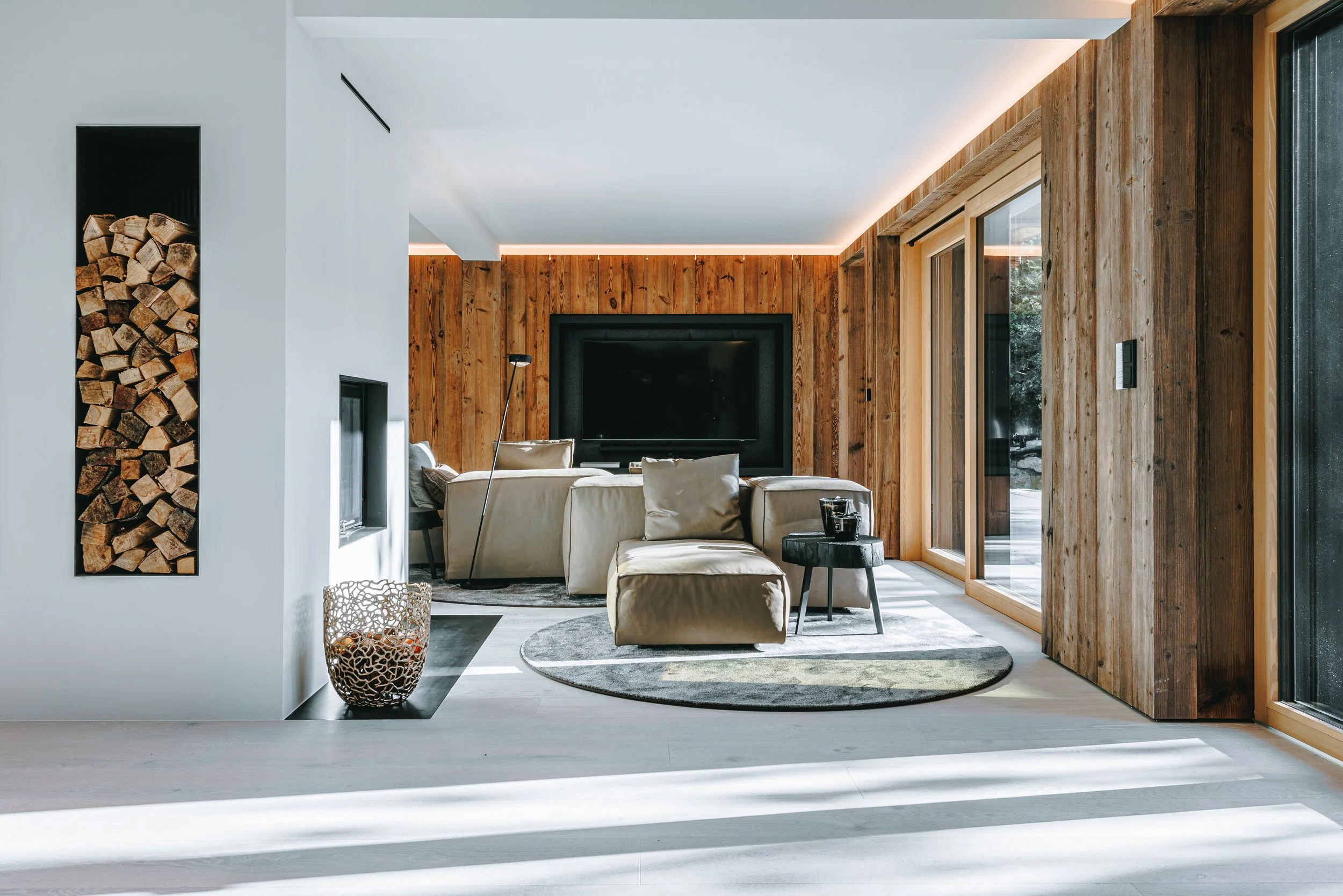 Modernes Wohnzimmer mit Holzverkleidung, großen Fensterfronten, beigen Sofas, Flachbildfernseher und Kamin mit Holzstämmen
