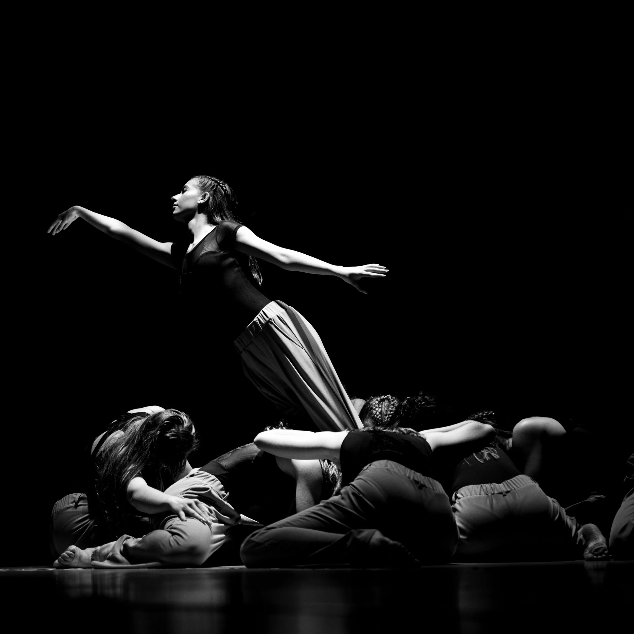 Eine Gruppe von Tänzern auf der Bühne. Eine Tänzerin befindet sich in der Mitte, die auf ausgestreckten Armen balanciert, während die anderen Tänzer sich kriechend im Halbkreis um sie herum befinden.