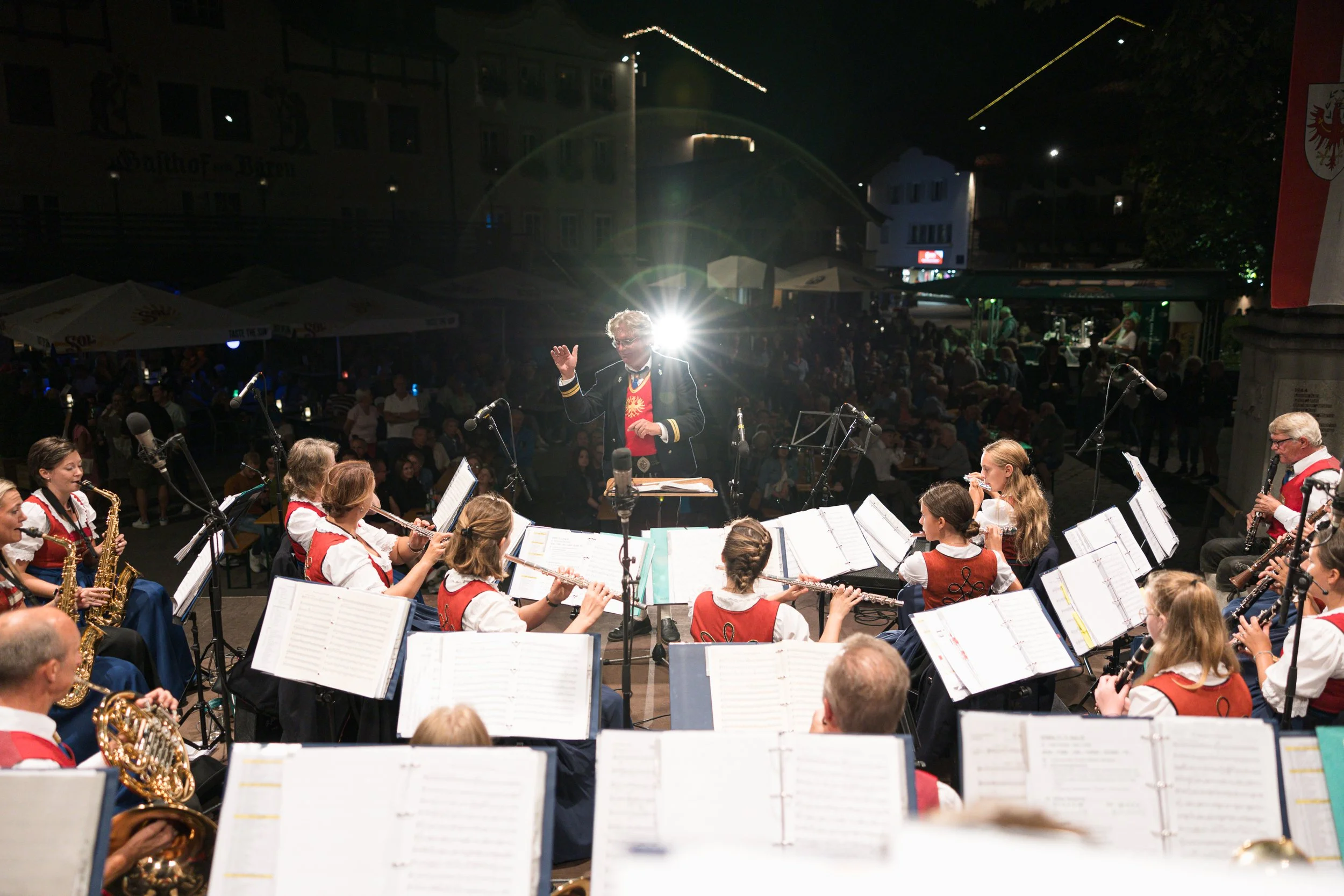 Eine große Orchestergruppe bei einem Auftritt auf einer Bühne im Freien bei Nacht, mit einem Dirigenten in der Mitte, der das Orchester leitet, während im Hintergrund das Publikum sitzt, draußen unter Zelten und Lichtern.