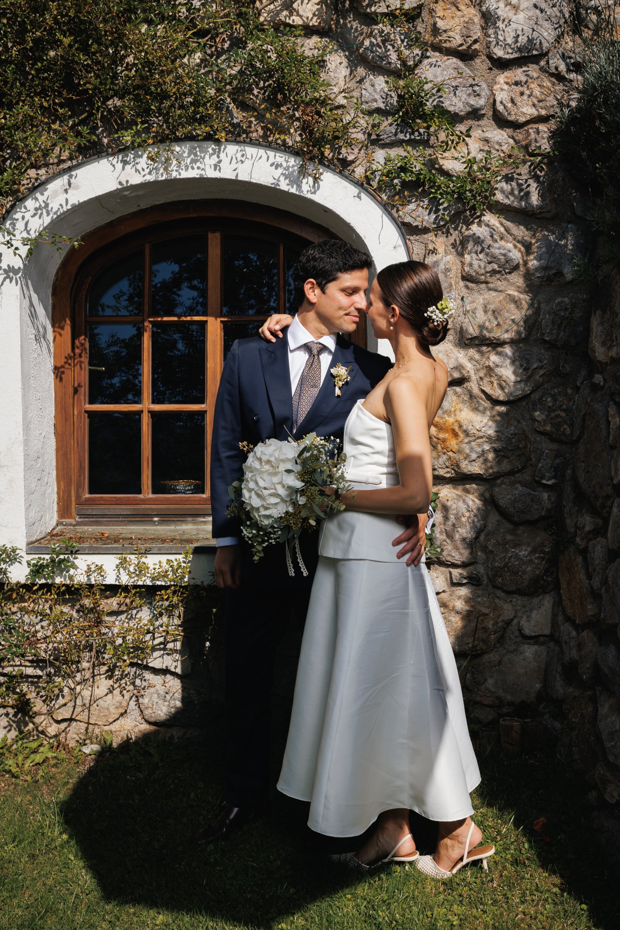 Ein Brautpaar steht sich romantisch vor einer Steinmauer mit einem kleinen Fenster, die Frau trägt ein weißes Brautkleid und hält einen Blumenstrauß, der Mann trägt einen Anzug, beide schauen sich verliebt an.