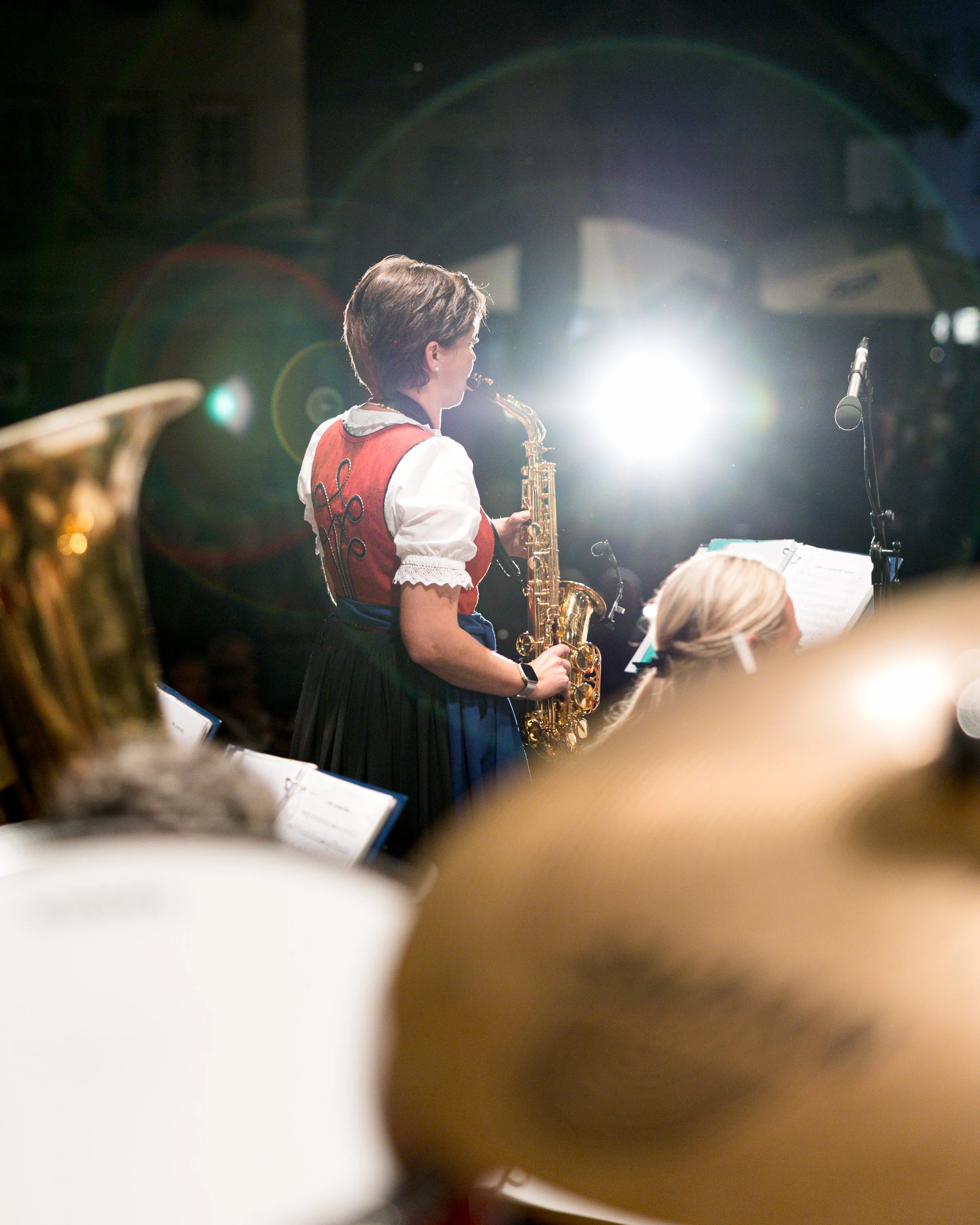 Eine Frau spielt auf einer Bühne Saxophon, getragen in traditioneller regionaler Kleidung, mit einem Mikrofon und Musiknoten im Vordergrund.