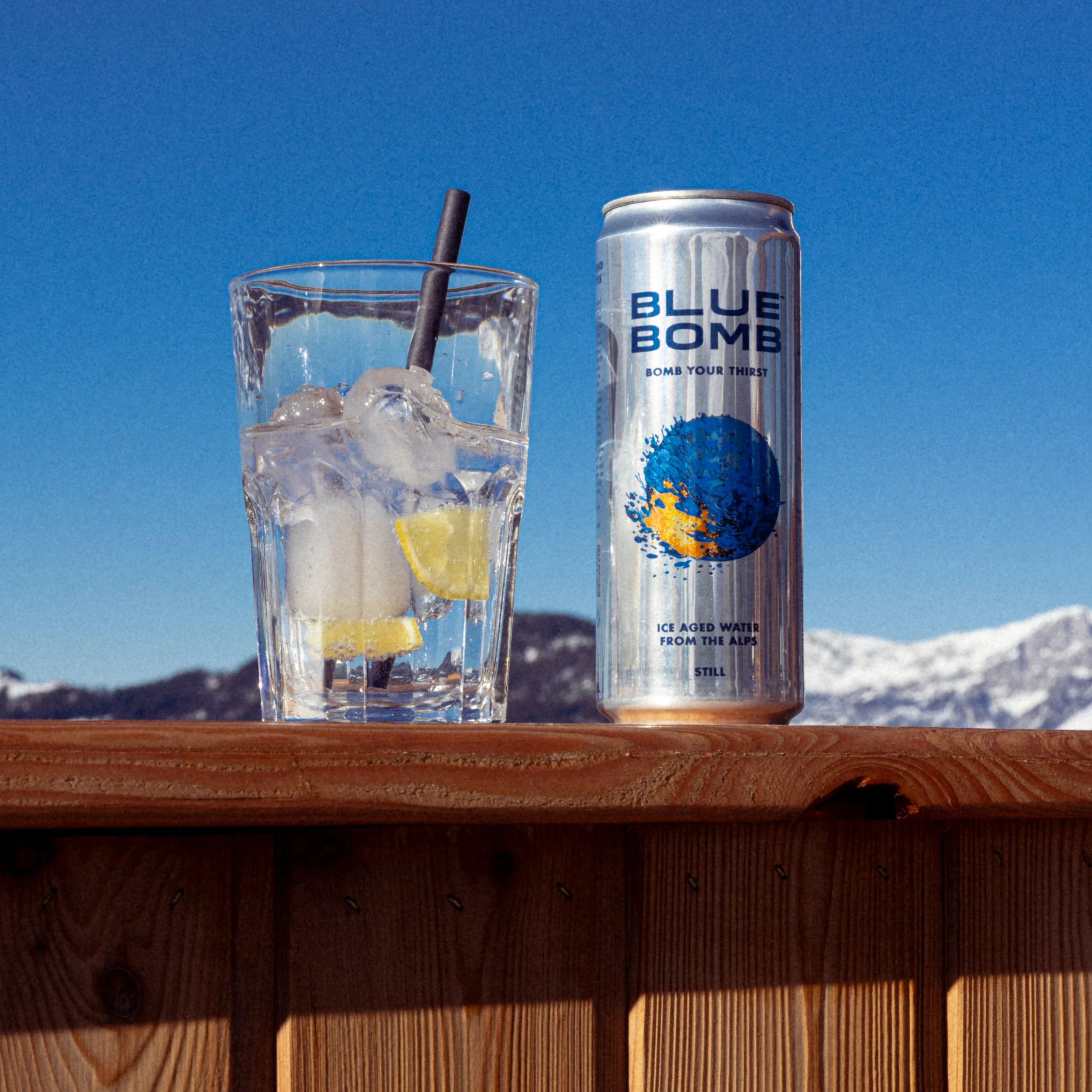 Ein Glas mit Eiswürfeln, Zitronenscheiben und einem schwarzen Strohhalm neben einer Dose mit dem Label 'Blue Bomb' vor einer Bergkulisse mit Schneegipfeln.