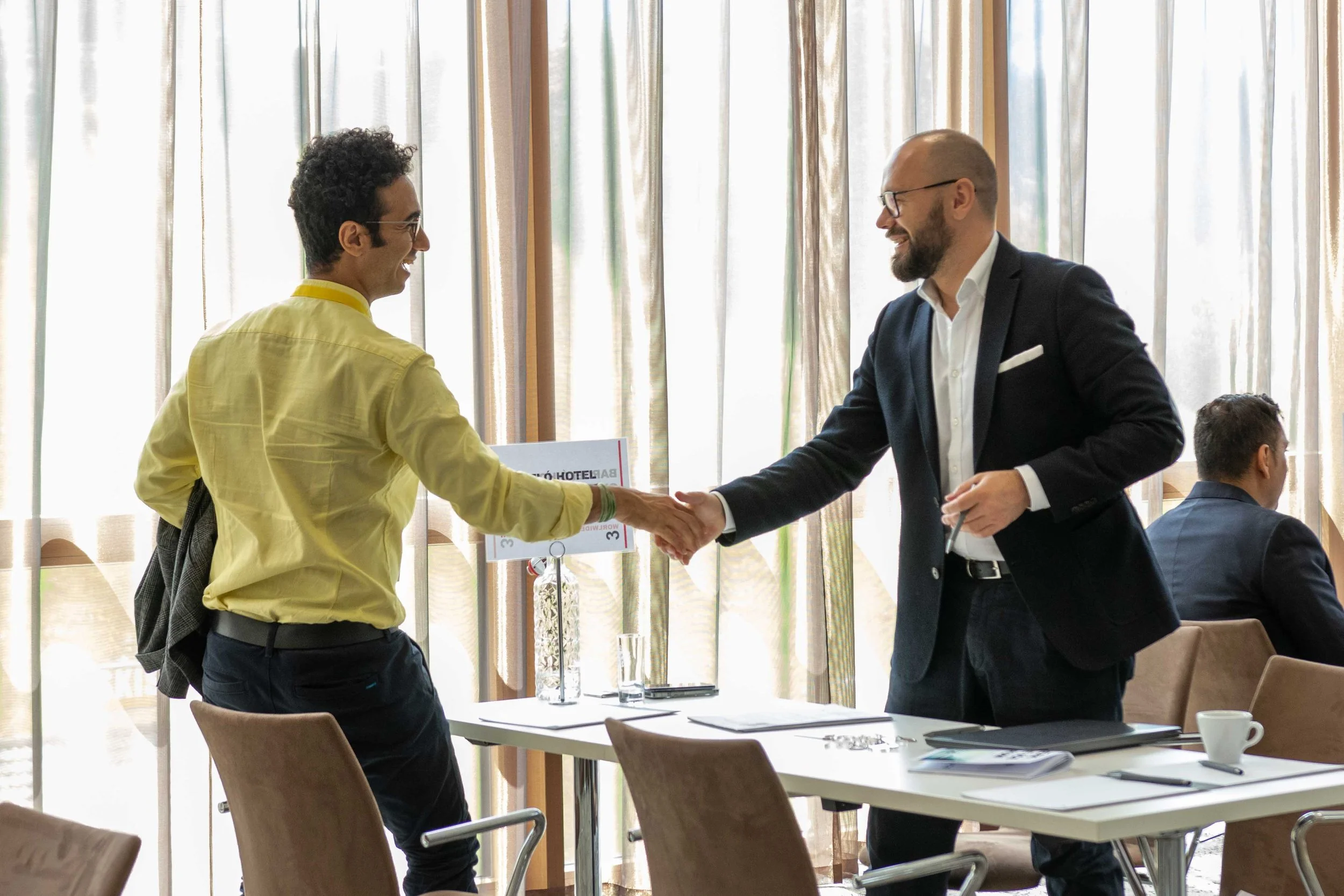 Zwei Geschäftsleute schütteln sich die Hand in einem modernen Meetingraum, während im Hintergrund andere Personen sitzen.