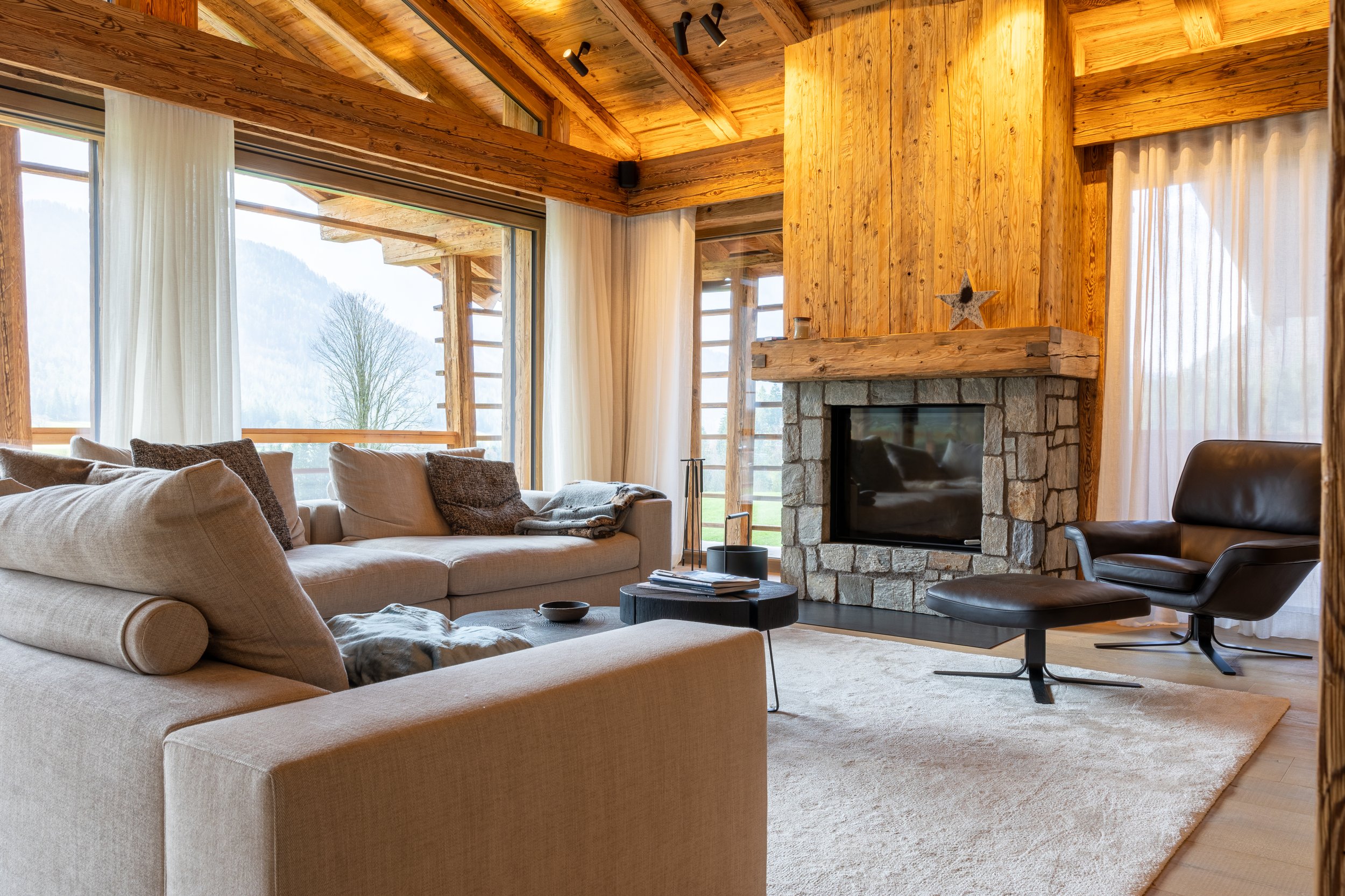 Gemütliches Wohnzimmer mit großen Fenstern, Sofa, Sessel, Kamin und Holzwänden im rustikalen Stil.