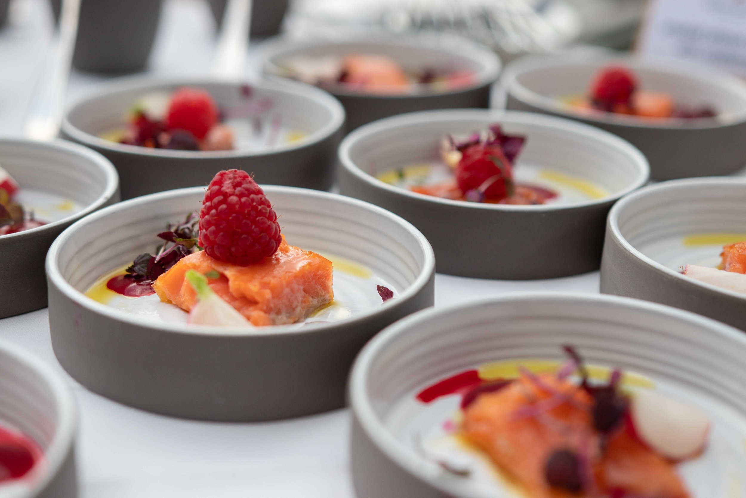 Mehrere kleine Schalen mit einem Gericht, das aus einer löffelförmigen Portion Lachs, einem roten Fruchtpüree, einem roten Beerenobst, einem weißen Saucen- oder Creme-Element und grünem Garnitur bestehen.