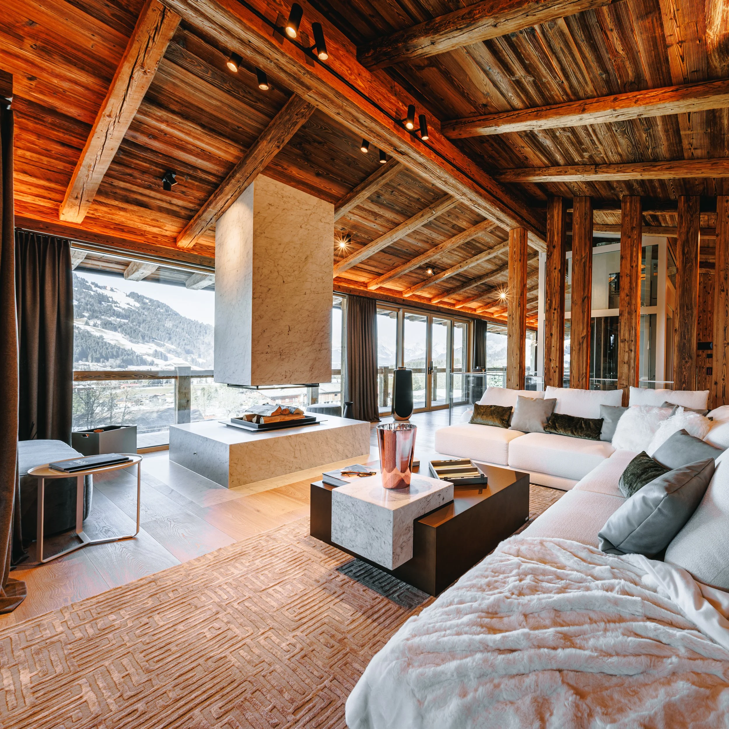 Ein modernes Wohnzimmer mit holzvertäfeltem Decke und Wänden, großen Fenstern mit Blick auf verschneite Berge, darunter ein weißes Sofa mit Kissen, einem schwarzen Couchtisch mit Büchern, einem Kamin aus Marmor in der Mitte des Raumes, und Vorhängen.