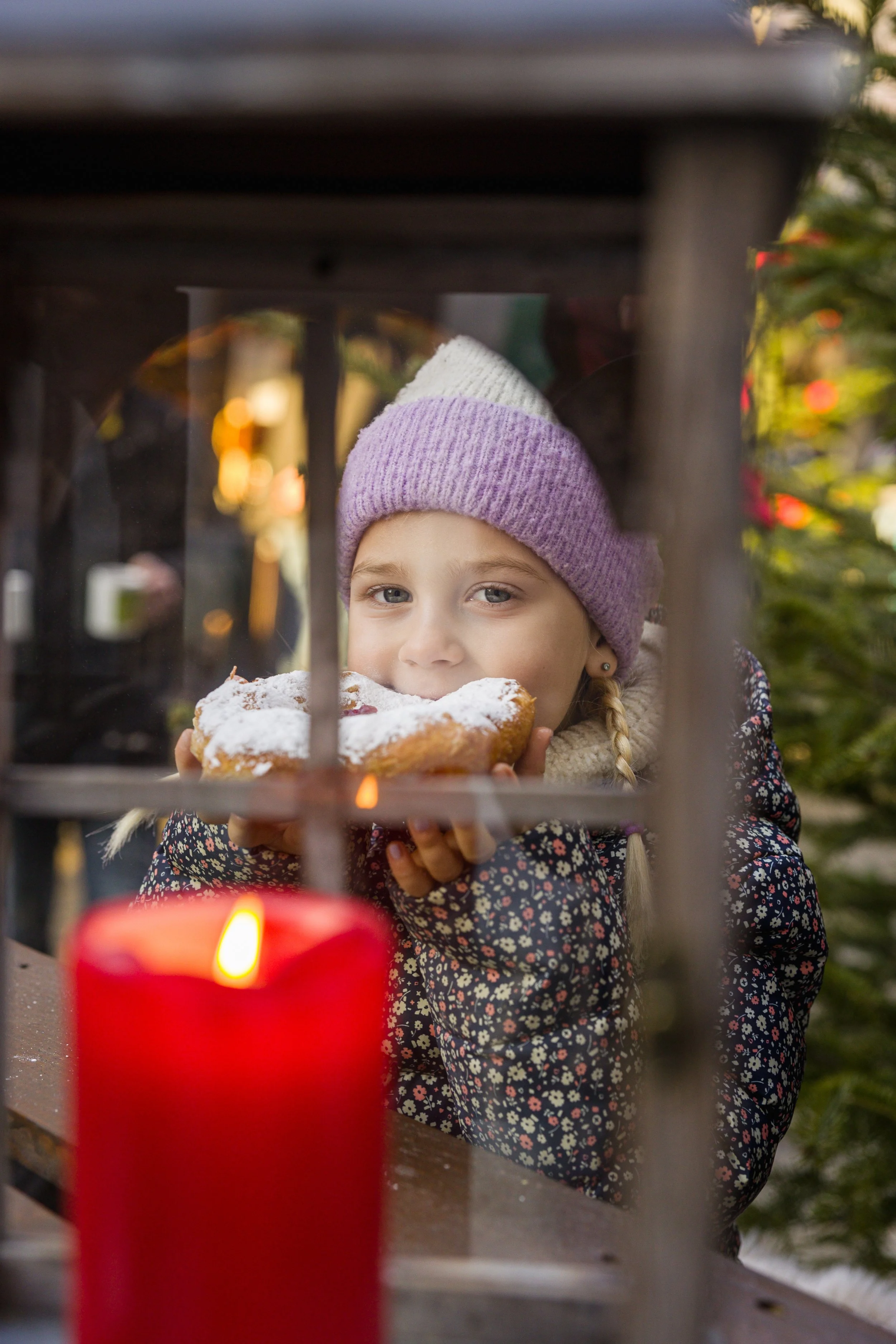 Ein junges Mädchen trägt einen lila und weißen Strickmütze und eine bunte Jacke und isst ein Stück Kuchen mit Puderzucker, während sie durch eine Fensteröffnung schaut, im Hintergrund leuchten Lichter und es sind Tannenbäume zu sehen.