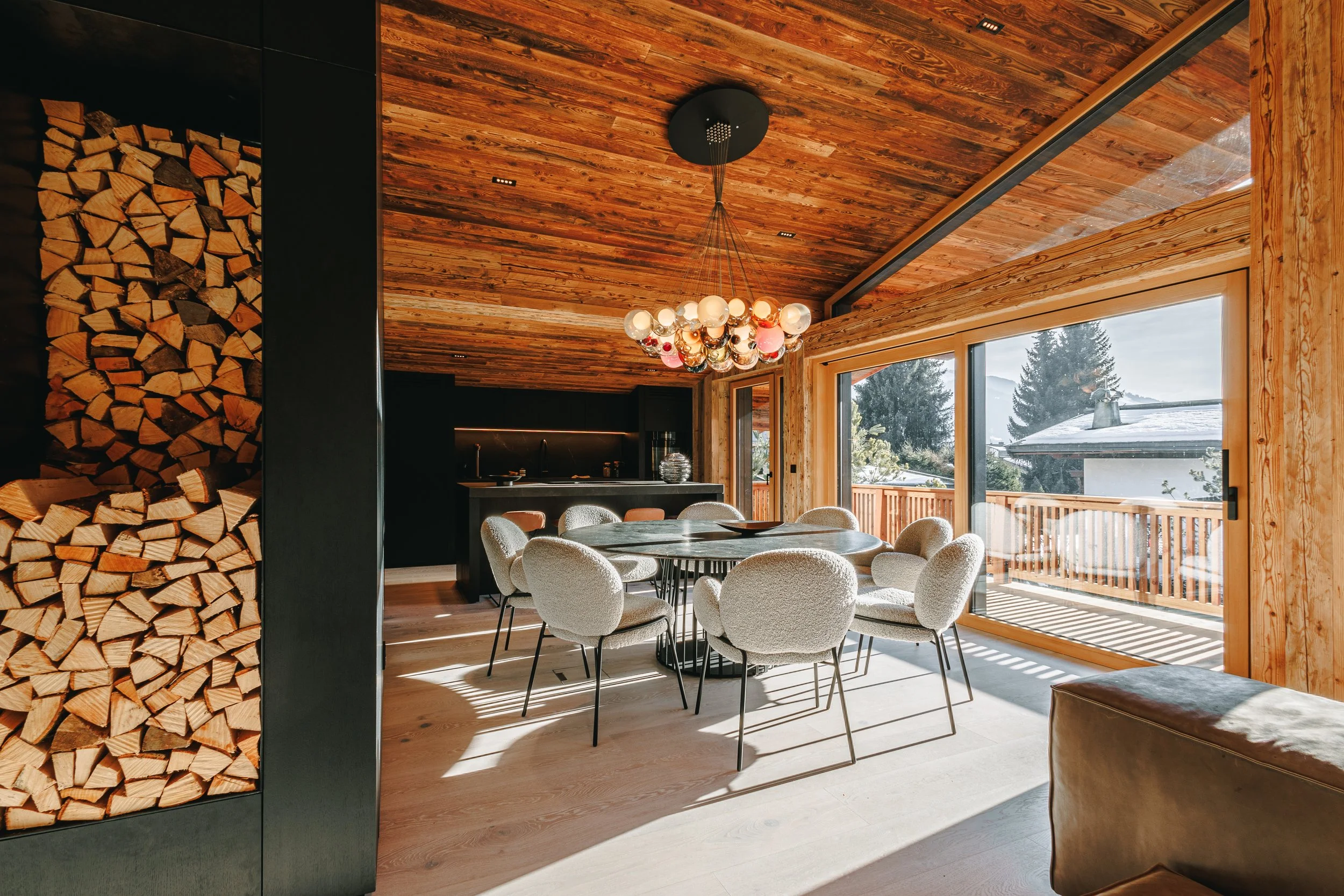 Moderner Essbereich in einem Chalet mit Holzdecken, großen Fenstern, einem runden Esstisch und bequemen Stühlen, mit Blick auf eine Schneelandschaft draußen.