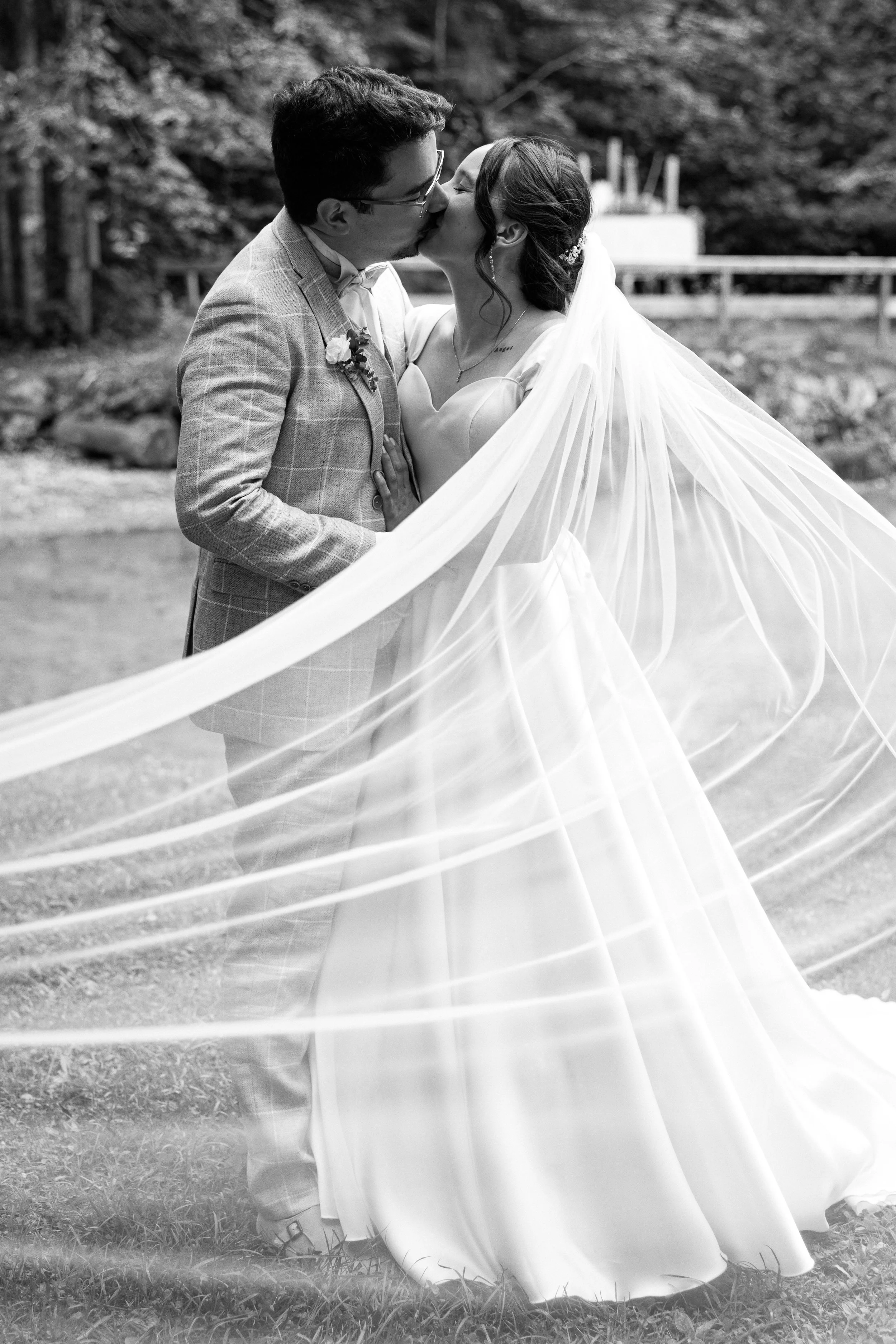 Ein Brautpaar bei ihrer Hochzeit, das sich küssend in einem Park umarmt, die Braut trägt ein langes, fließendes Kleid und ein Schleier, der im Wind weht.