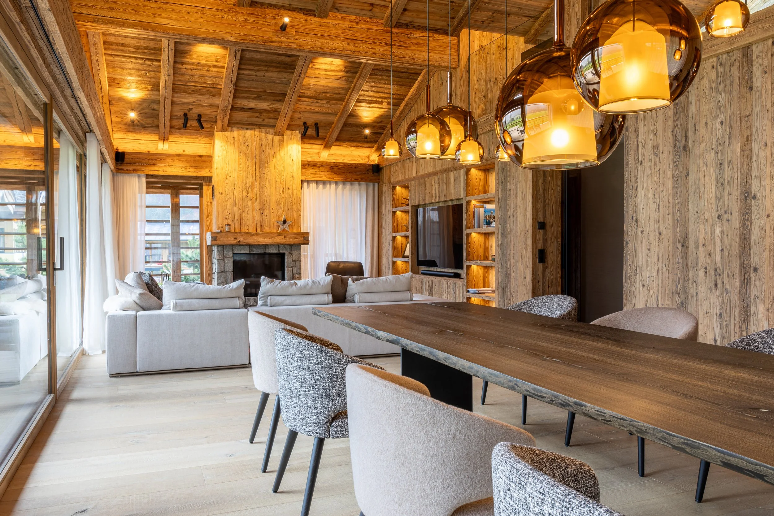 Innenraum eines Wohnzimmers mit Holzdecken, Kamin, Sofa, Esstisch und modernen Leuchtlampen