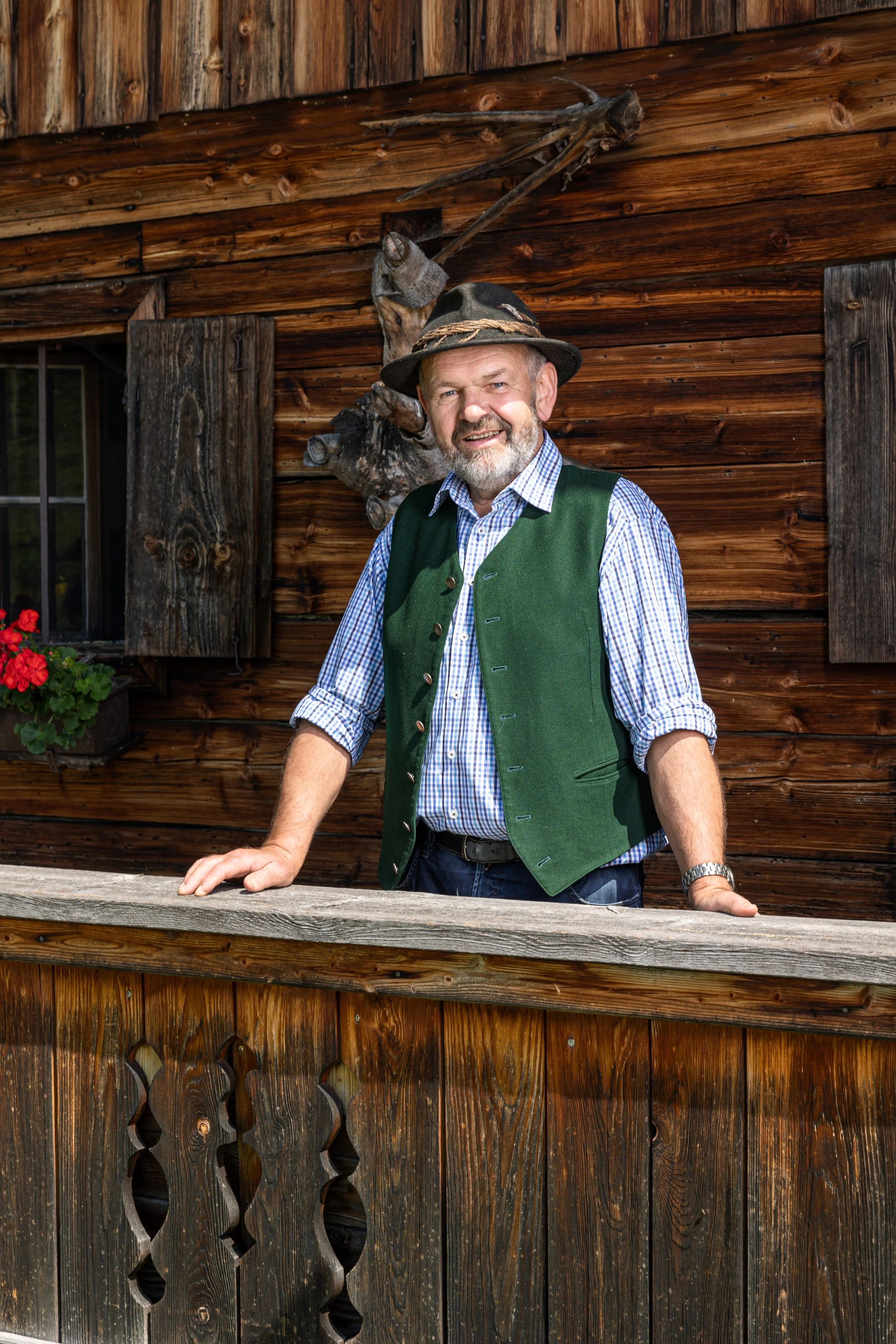 Ein älterer Mann in traditioneller bayerischer Kleidung mit grüner Weste und kariertem Hemd, der auf einer Holzveranda steht, vor einem Holzhaus mit Holztüren und -fenstern, einem Fensterpflanzenkasten mit roten Blumen und einem Hirschgeweih an der W