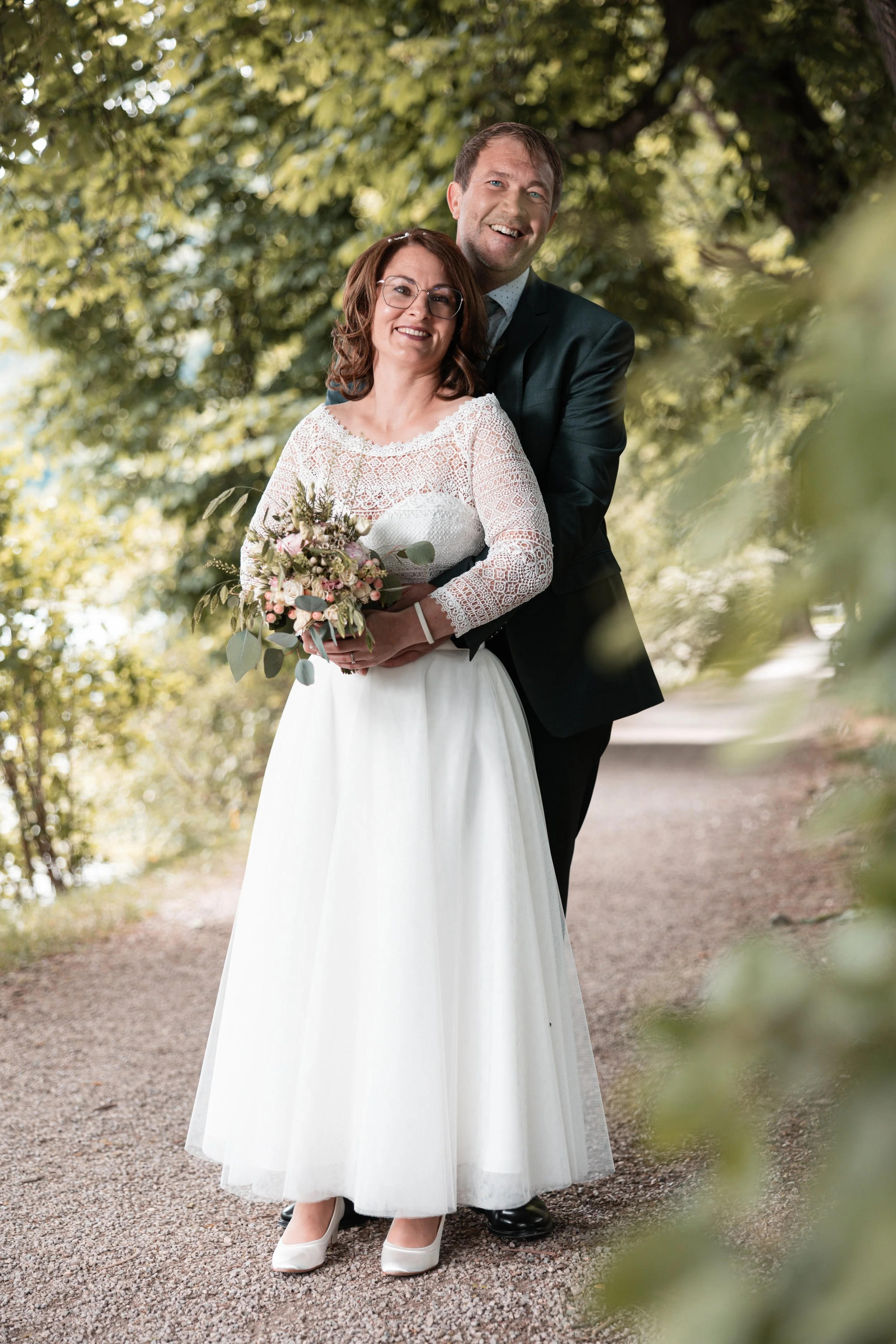 Ein lächelndes Brautpaar steht auf einem Waldweg, die Braut trägt ein weißes Kleid mit Spitze und hält einen Blumenstrauß, der Bräutigam trägt einen dunklen Anzug.